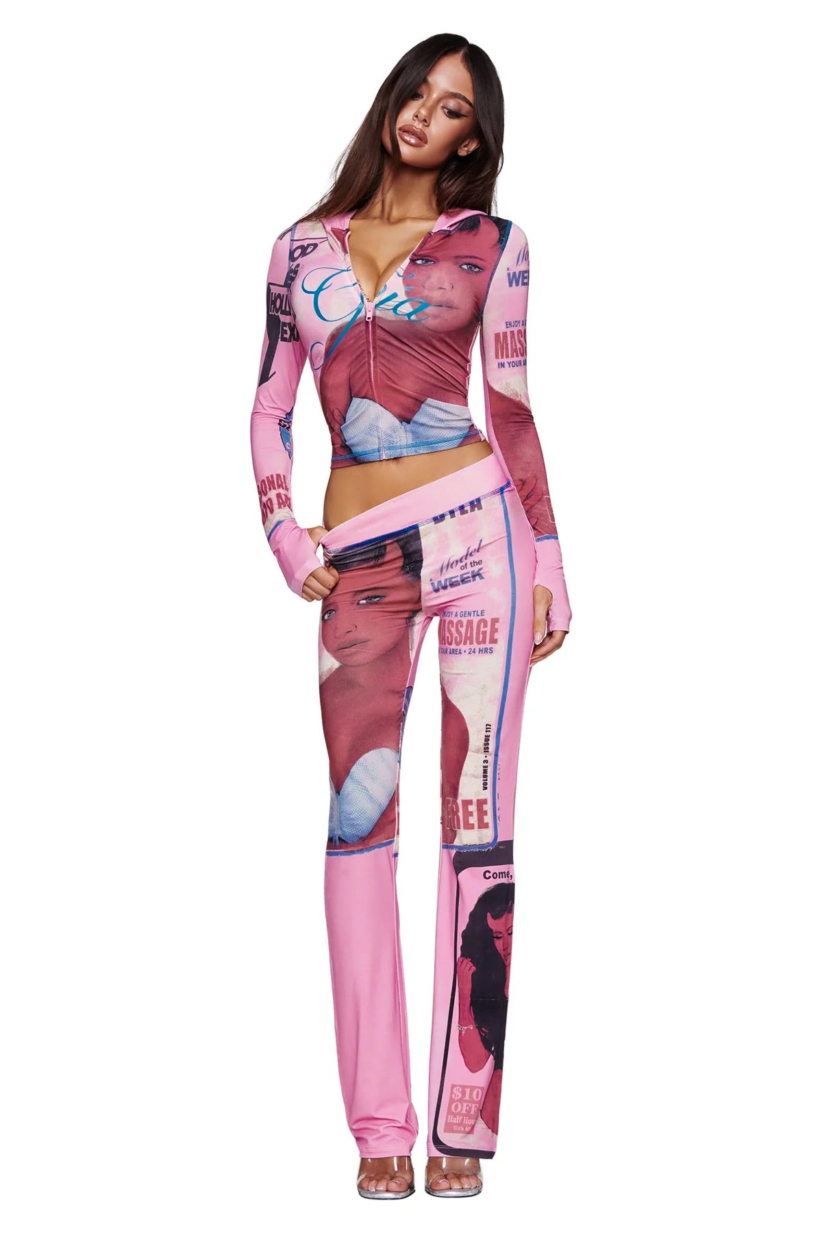A woman stands facing forward with one hand on her hip and long, straight hair, wearing the I.AM.GIA T-REX BLARE HOODIE in pink. The hoodie features bold vintage magazine cover prints with female faces and text, channeling the brand's signature streetwear attitude.