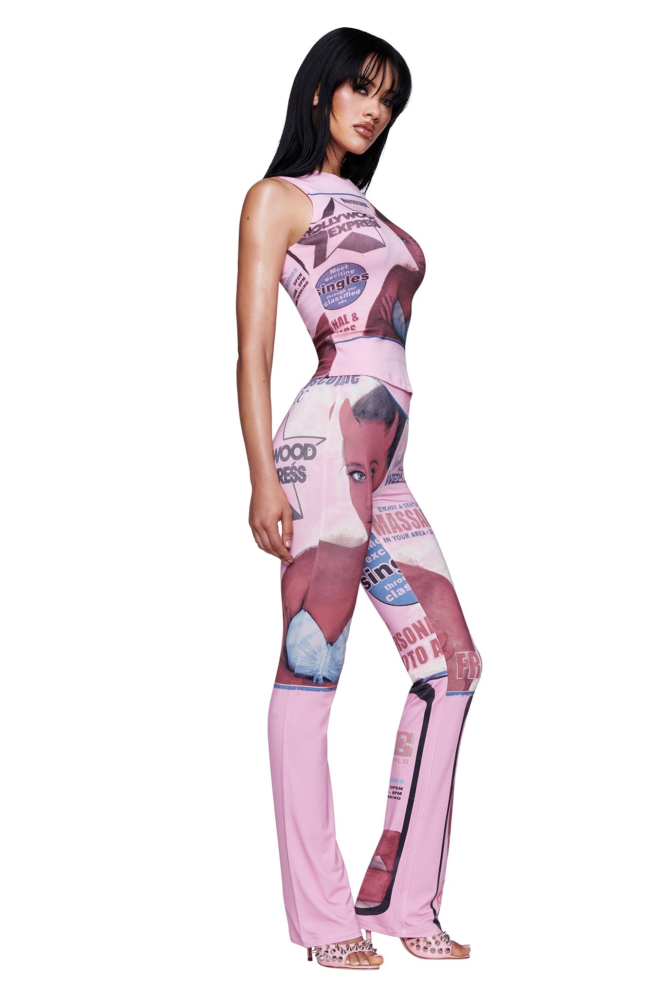 A woman with long dark hair stands in profile against a white background, wearing the I.AM.GIA T-REX BLARE TRACKPANT in pink—fitted pants printed with bold magazine covers and images.