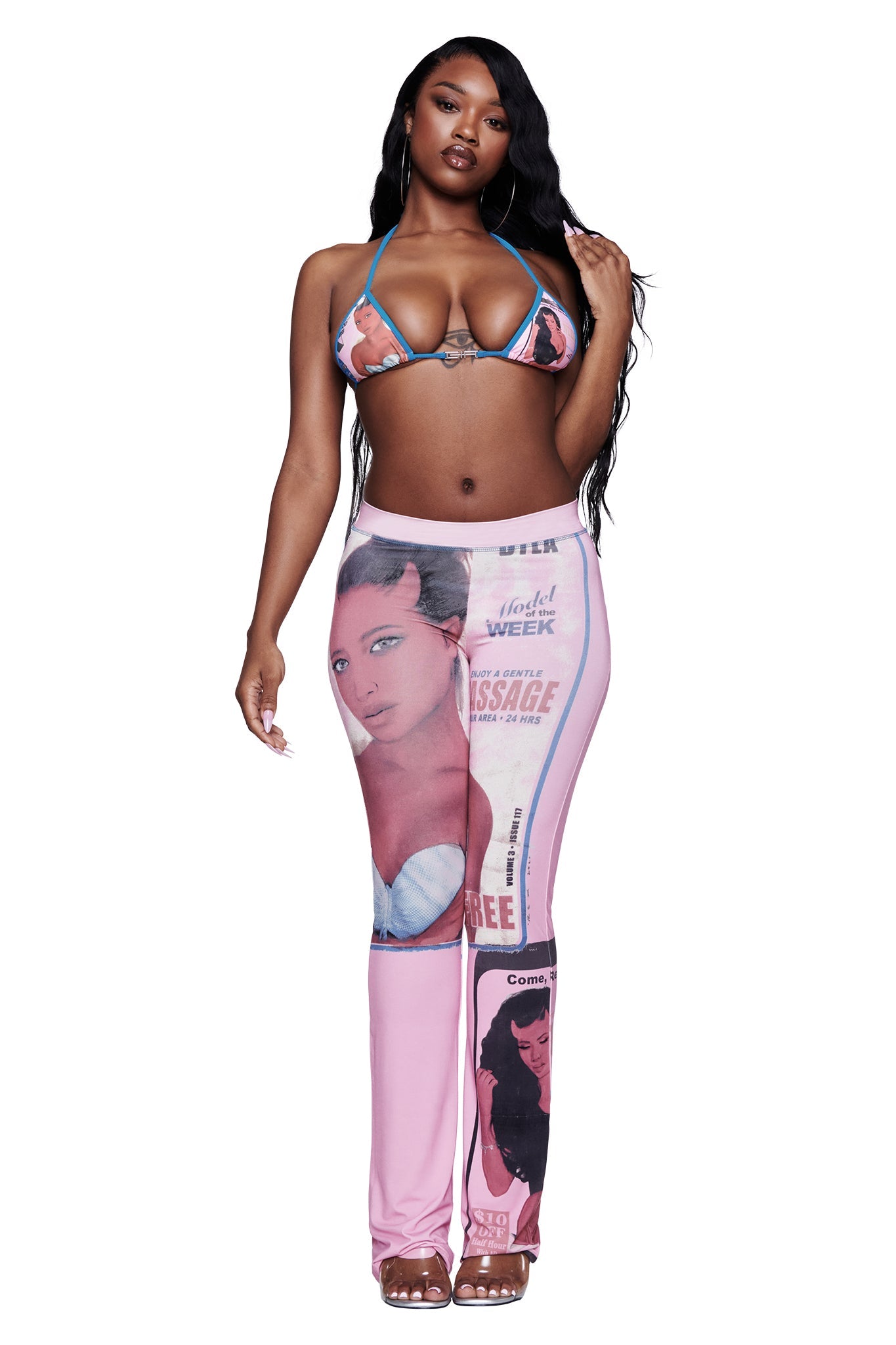 A woman in a bikini top and I.AM.GIA T-REX BLARE TRACKPANT - PINK, featuring bold magazine-style prints, stands against a white background, looking at the camera with one hand touching her hair.