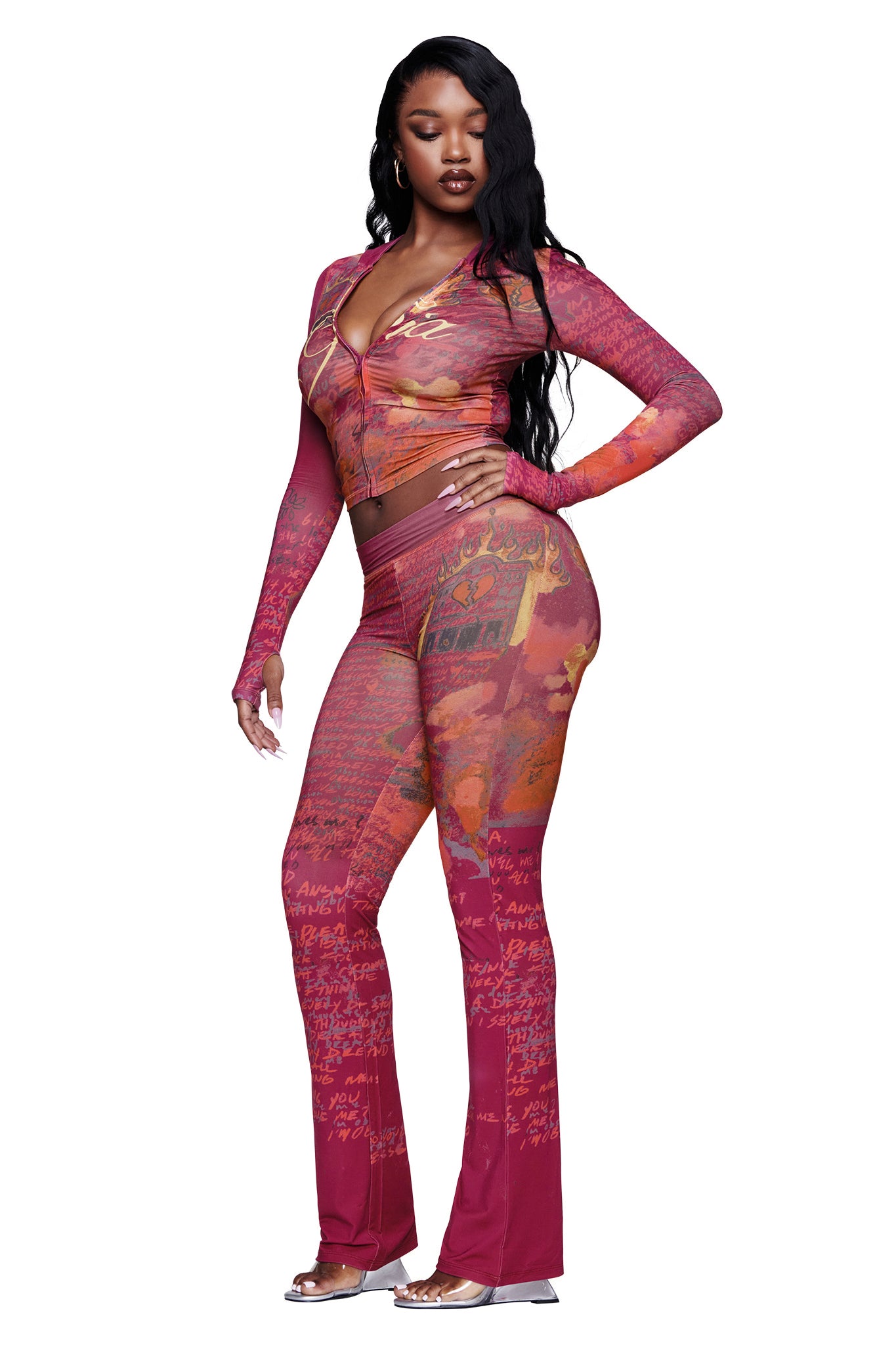 A woman stands against a white background with one hand on her hip, dressed in a fitted, long-sleeve sheer pink and orange patterned top paired with the I.AM.GIA T-REX BLARE TRACKPANT in red and open-toe heels.