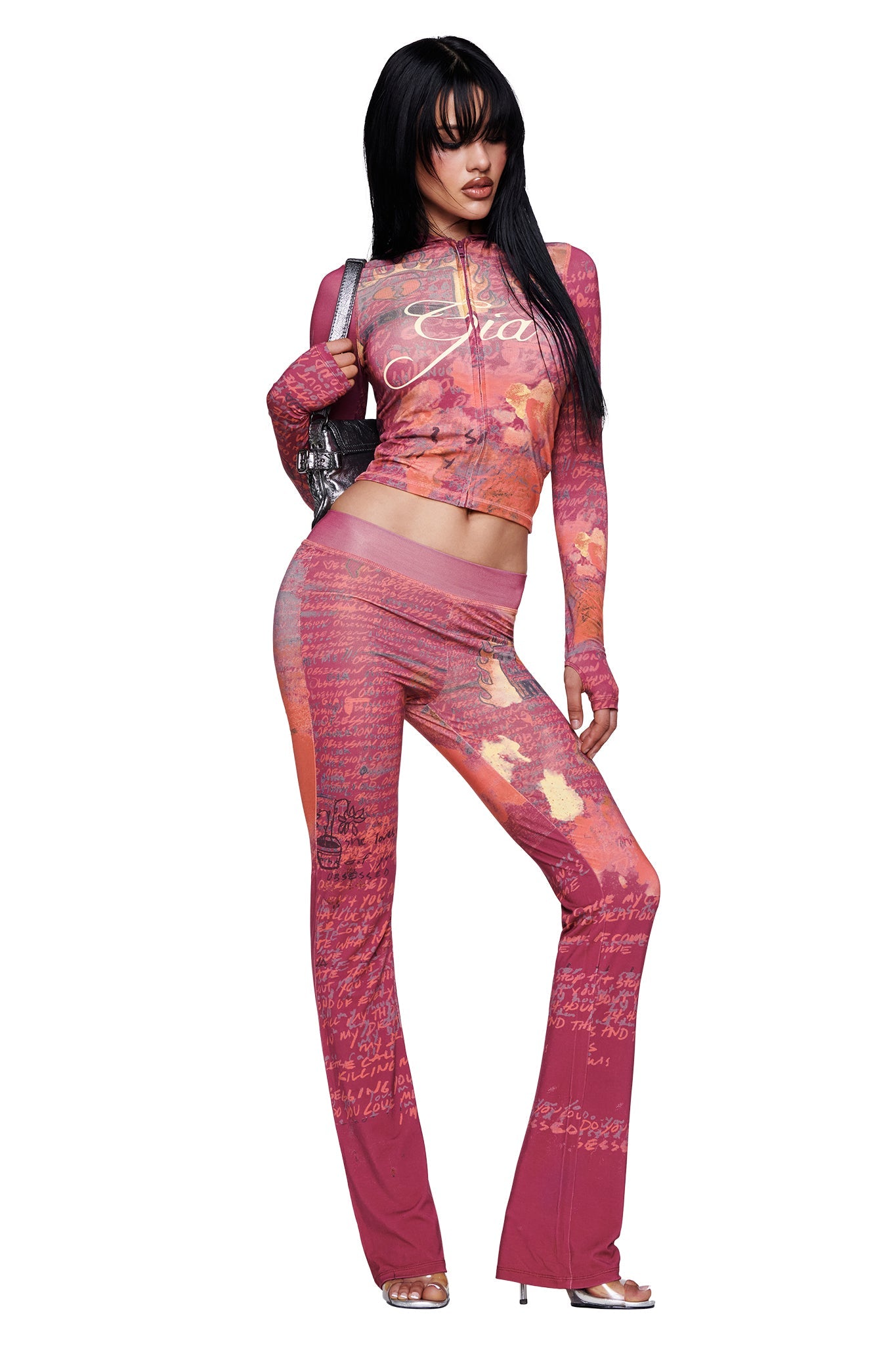 A person poses confidently in a bold, red, patterned two-piece ensemble with long sleeves and flared women's lounge pants inspired by the I.AM.GIA T-REX BLARE TRACKPANT - RED, accessorized with a small silver handbag, heeled sandals, and styled with long dark hair and bangs.