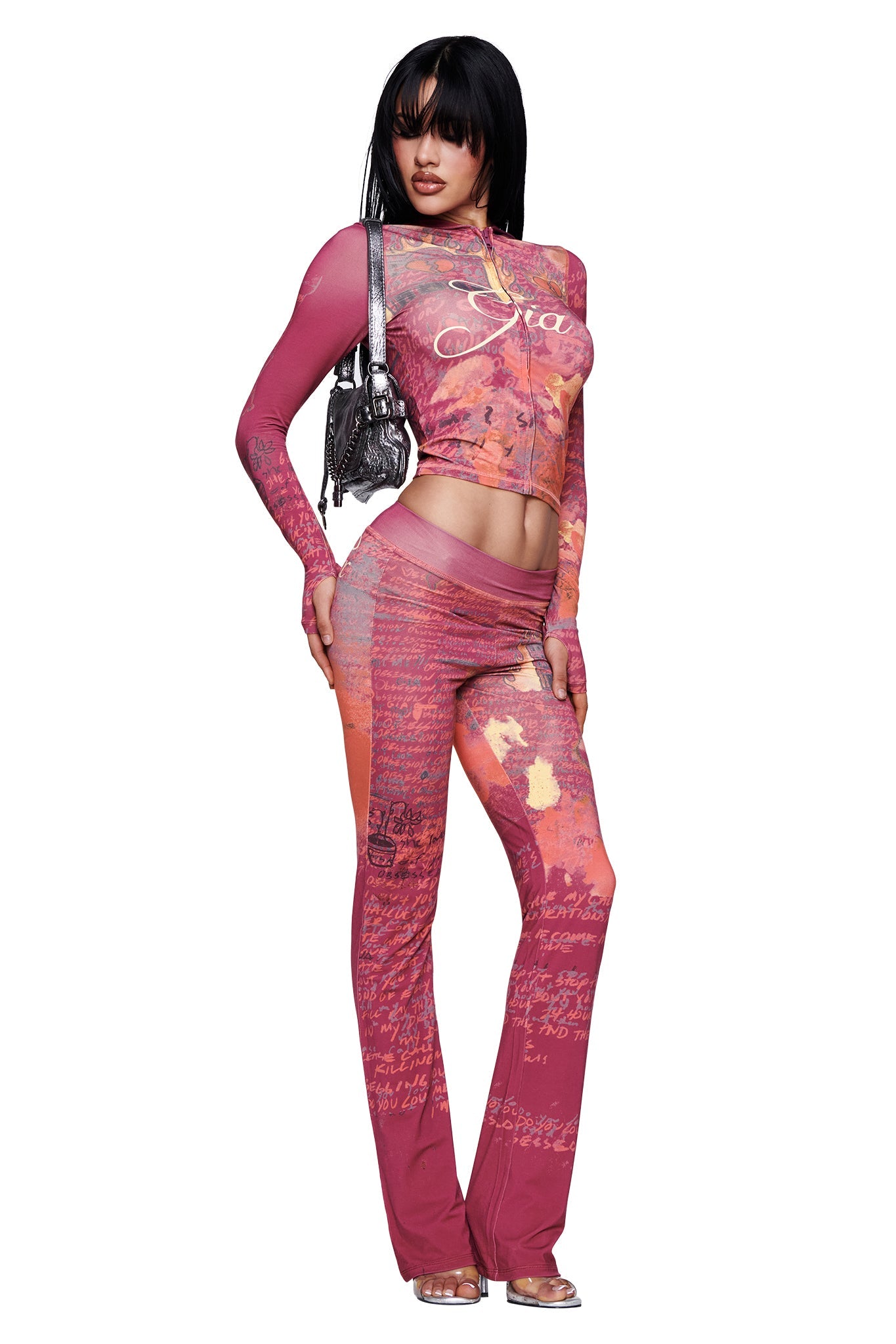A person poses in the I.AM.GIA T-REX BLARE TRACKPANT - RED, styled with high heels and a small silver purse against a white background. The bold red print of the trackpant adds an extra pop of color to the outfit, making it a standout look.