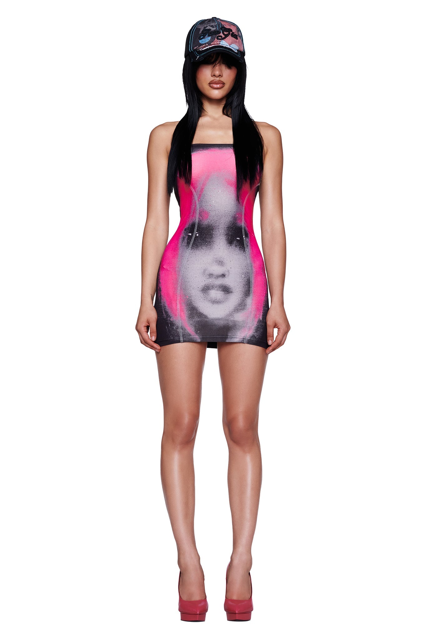 A woman wearing the I.AM.GIA T-REX BLARE BOOB TUBE MINI DRESS in black stands confidently against a white background, styled with pink platform heels and a black baseball cap.