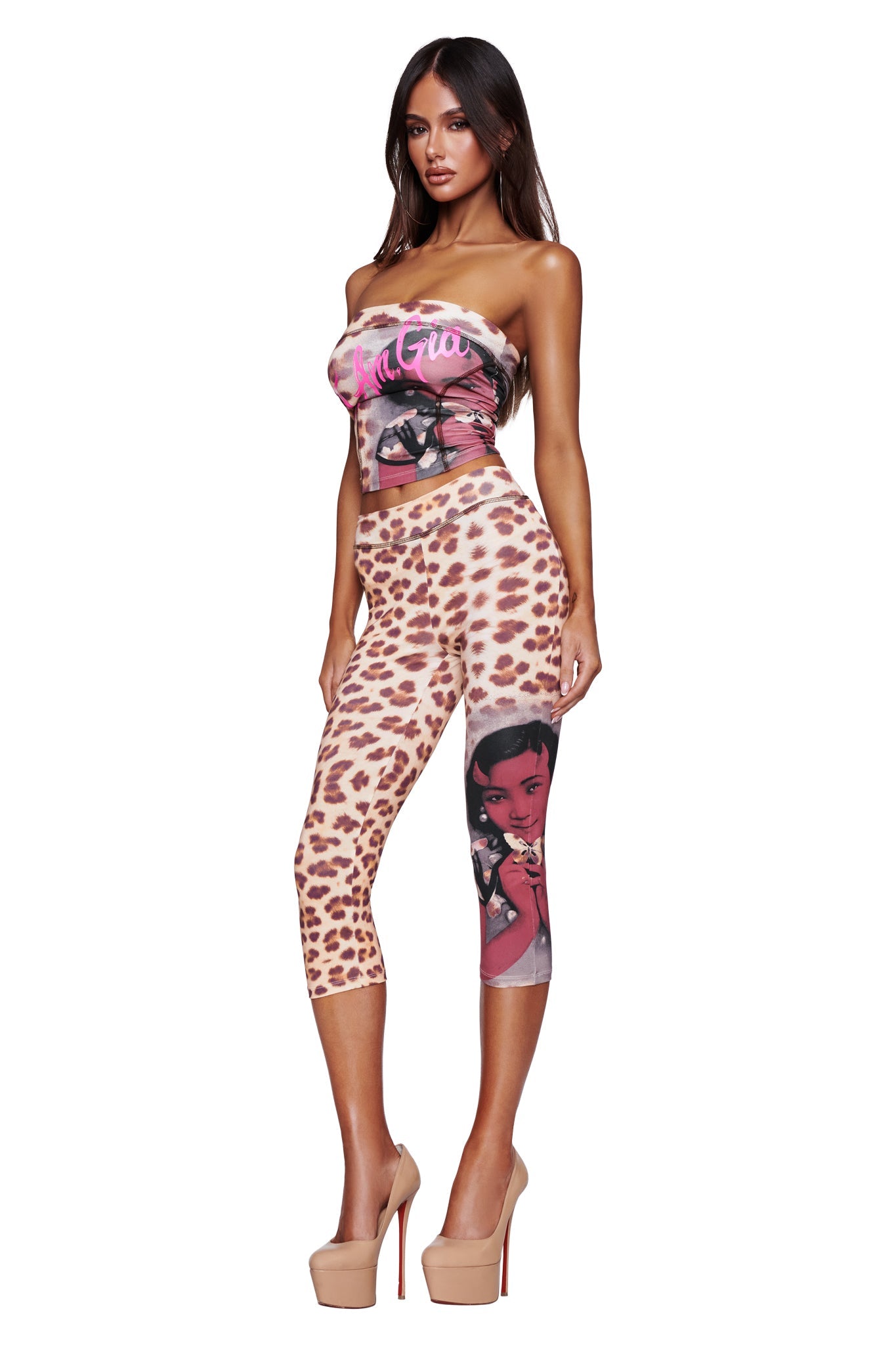 A woman wearing the I.AM.GIA T-REX BLARE CAPRI - WILD CARD, which features a cheetah print and a cartoon graphic on one leg, paired with a strapless top adorned with text and graphics, and nude high heels, standing against a white background.