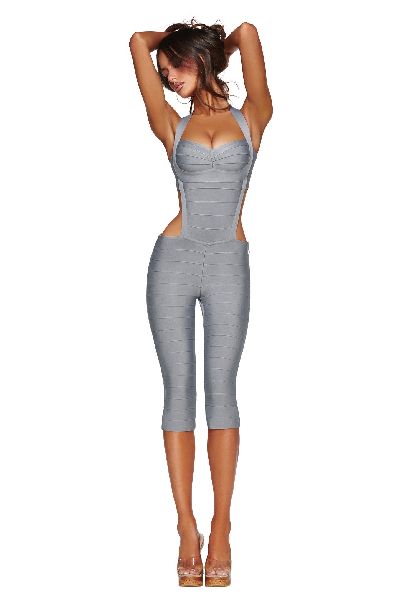 Model wears the I.AM.GIA EVA BANDAGE CAPRI JUMPSUIT in grey—a form-fitting, sleeveless catsuit featuring cutout sides—paired with clear high heels, posing with arms raised above her head against a plain white background.