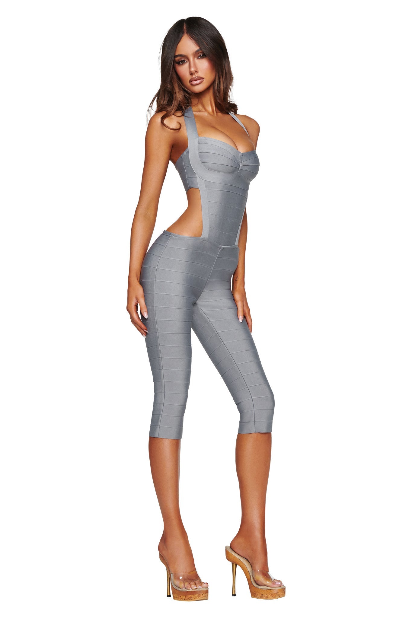 A woman poses in the I.AM.GIA EVA BANDAGE CAPRI JUMPSUIT in grey—a fitted, sleeveless halter-neck style with cut-out sides—paired with clear high heels against a plain white background.