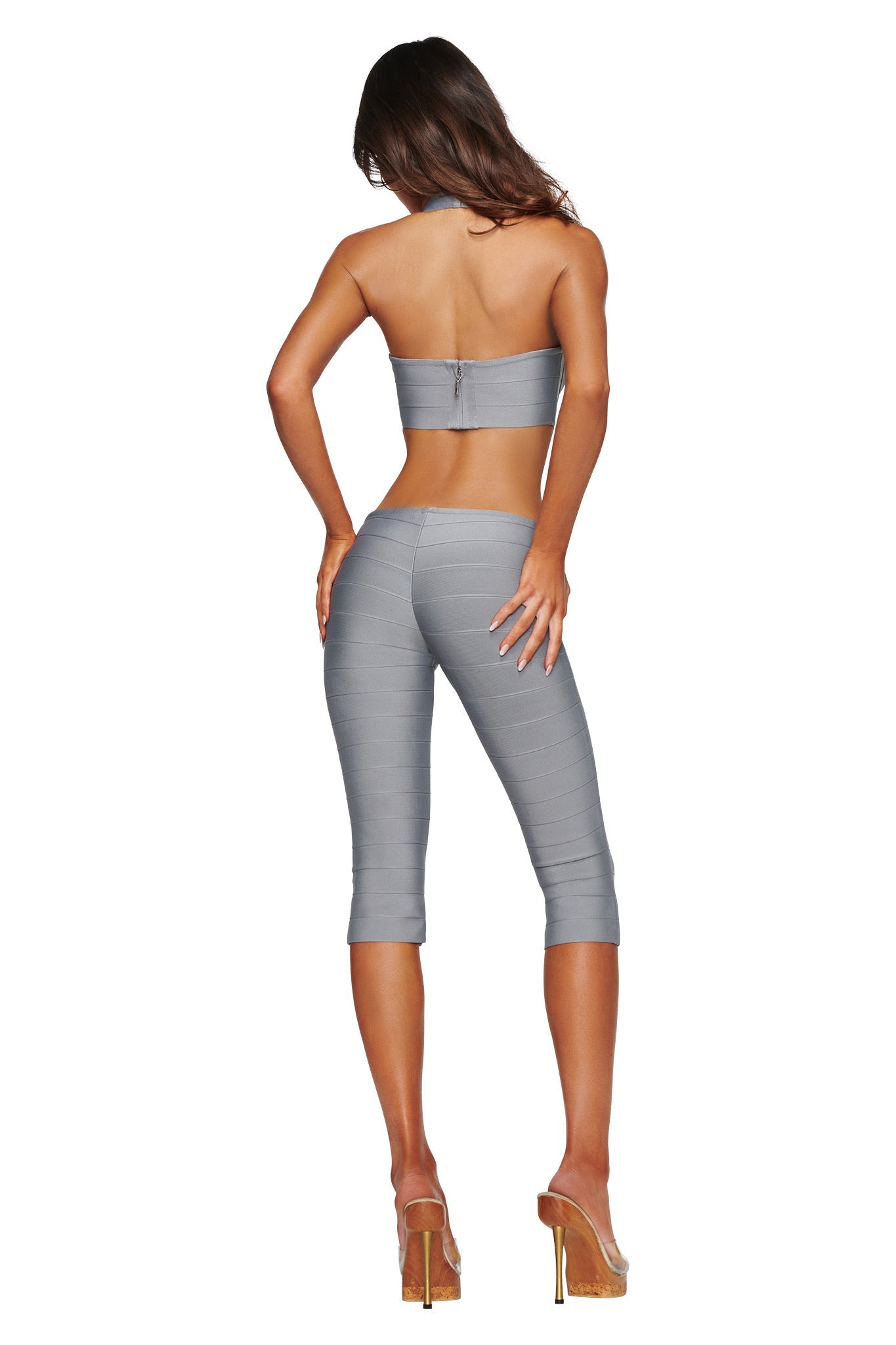 A woman stands facing away, wearing the I.AM.GIA EVA BANDAGE CAPRI JUMPSUIT in grey—a strapless bodycon piece that accentuates her silhouette—paired with clear high heels.