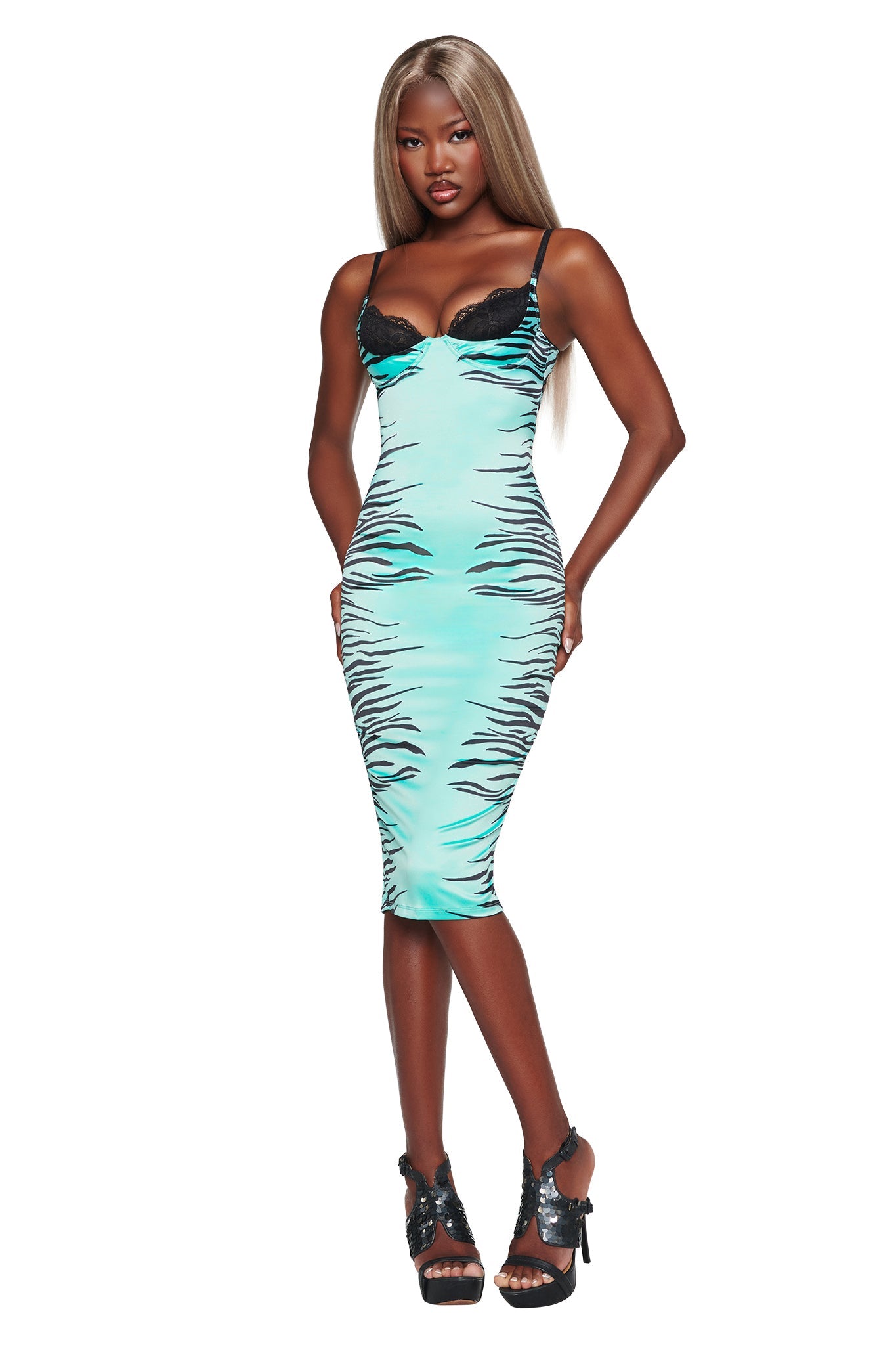A woman wearing the I.AM.GIA CASSY MIDI DRESS in aqua fine line zebra—a fitted bodycon silhouette featuring black lace cups—poses with hands on hips. She has long, straight hair and pairs the look with black high-heeled sandals against a white background.