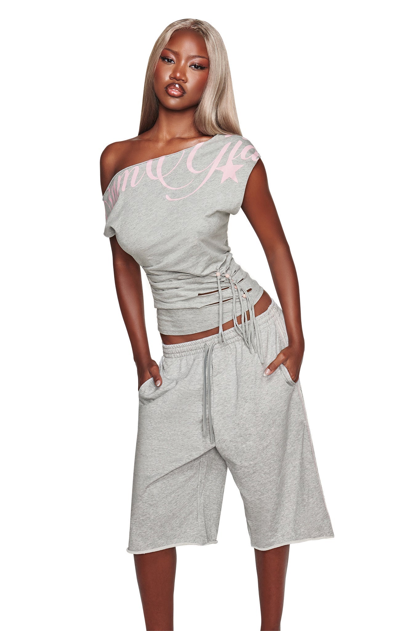 A woman stands confidently with her hands in her pockets, wearing the I.AM.GIA BEX TERRY SHORT in grey, styled with a light gray off-shoulder top accented by pink lettering and an elasticated waistband for a relaxed fit.