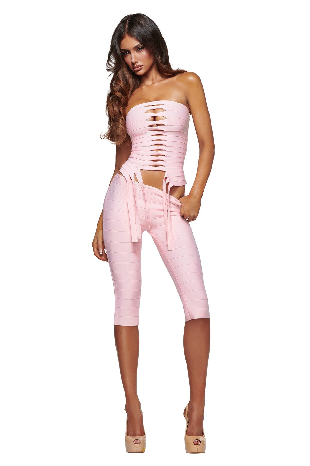 A woman models the I.AM.GIA MAEVE BANDAGE CAPRI - BABY PINK, a form-fitting outfit featuring cutouts on the top and capri-length bottoms, paired with heels and posing with one hand in her pocket against a white background.
