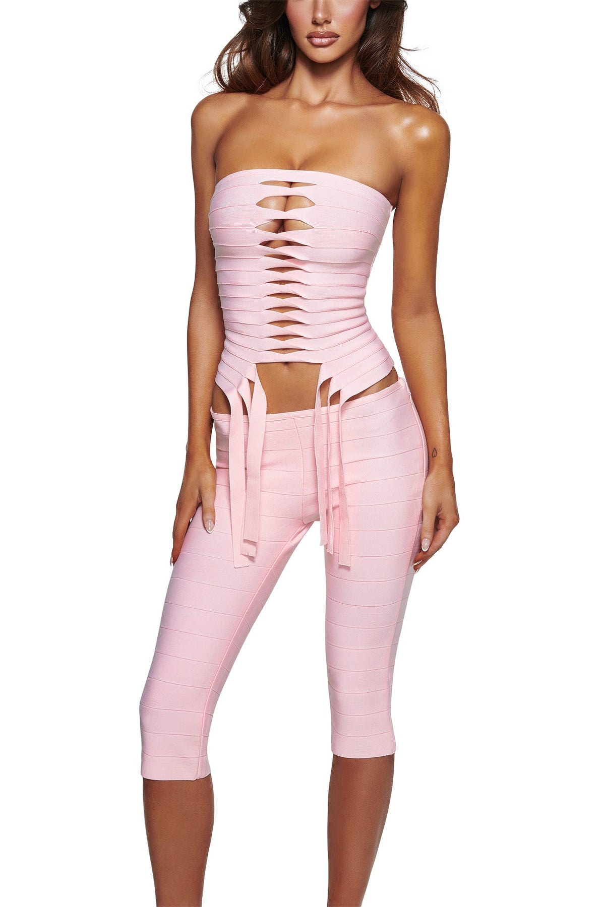 A woman wears the MAEVE BANDAGE CAPRI in baby pink by I.AM.GIA—a strapless ensemble with multiple front cutouts, horizontal slits on the top, ribbon-like ties at the waist, and knee-length bandage capri pants.