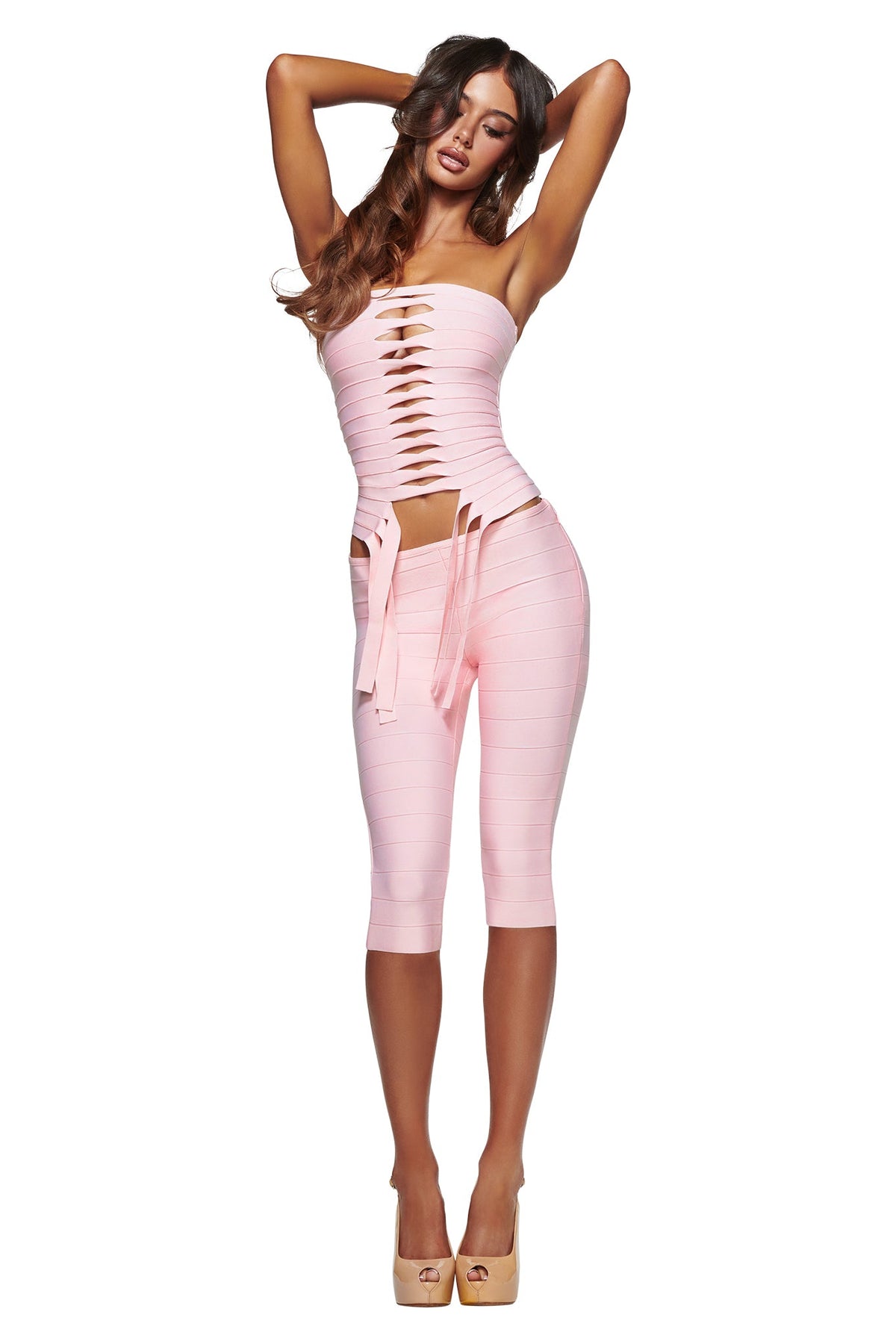 A woman is wearing the I.AM.GIA MAEVE BANDAGE CAPRI in baby pink, styled with a light pink cut-out strapless top and nude high heels, posing with her arms raised above her head.