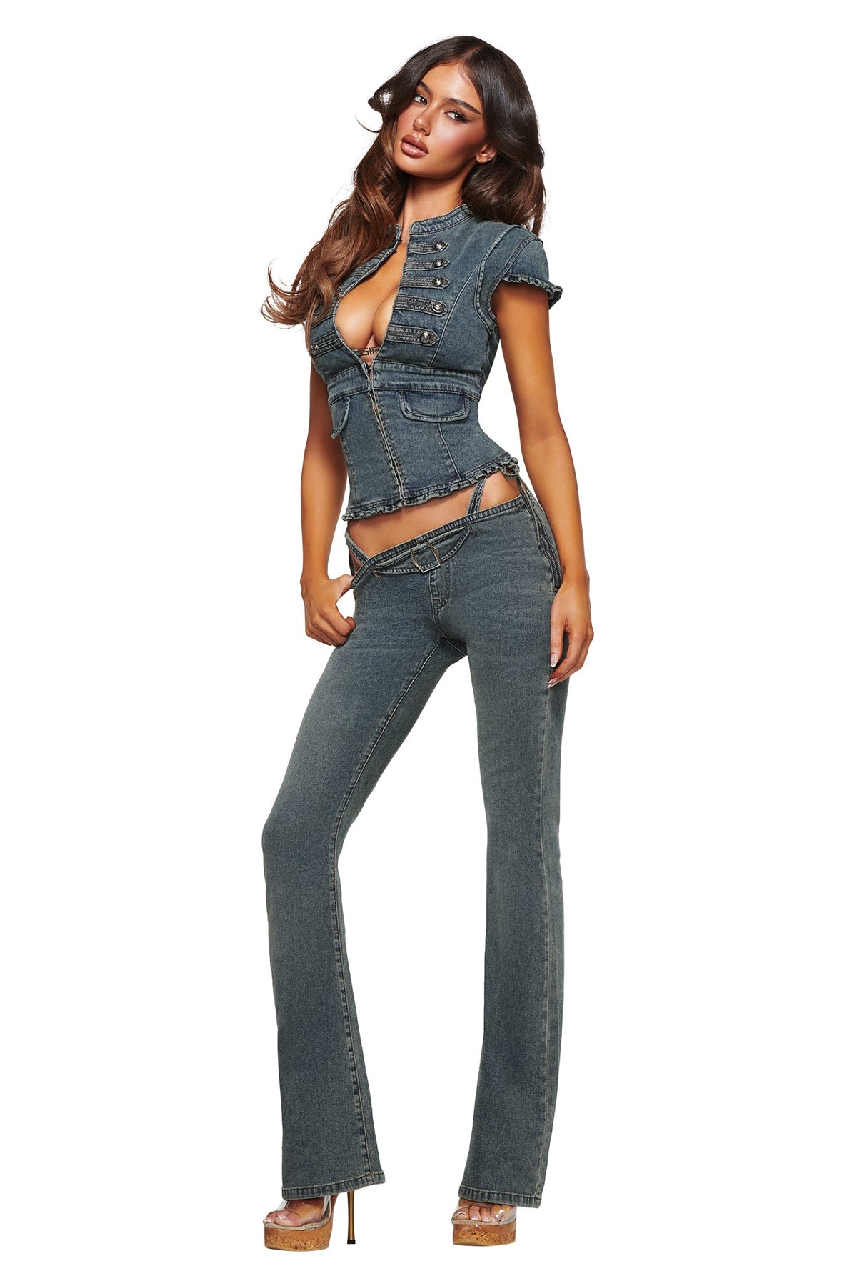 A woman models the I.AM.GIA JET G-STRING DENIM PANT in faded indigo, featuring a fitted silhouette with a deep neckline, exposed midriff, and low-rise flared jeans highlighted by a subtle G-string detail. She pairs the bold, Y2K-inspired look with high-heeled sandals and stands against a white background.