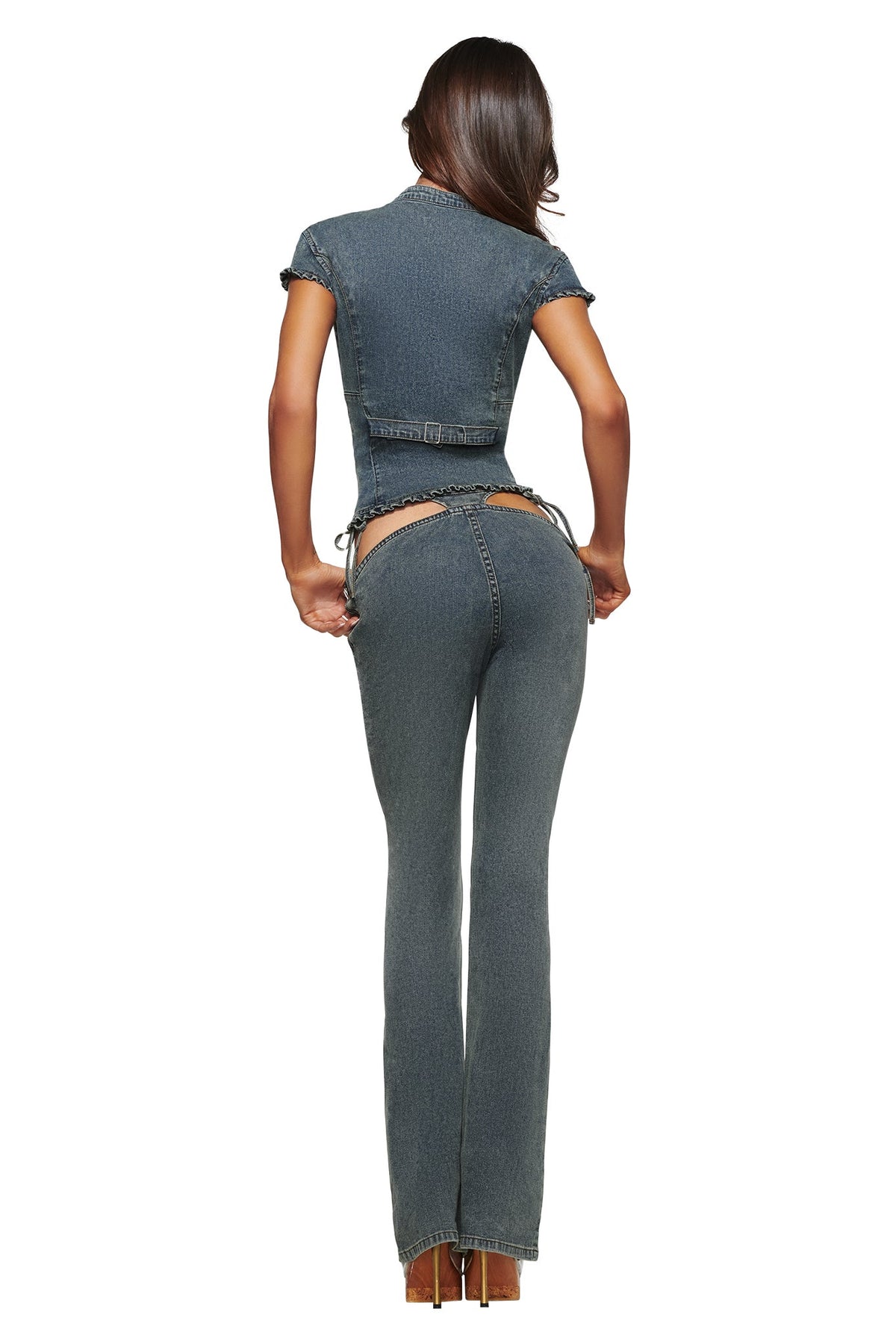 A woman stands facing away, wearing the I.AM.GIA JET DENIM TOP - FADED INDIGO with high-waisted denim pants featuring cutouts that reveal part of her lower back and upper thighs. She has long brown hair and wears high heels, channeling bold Y2K outfit vibes.