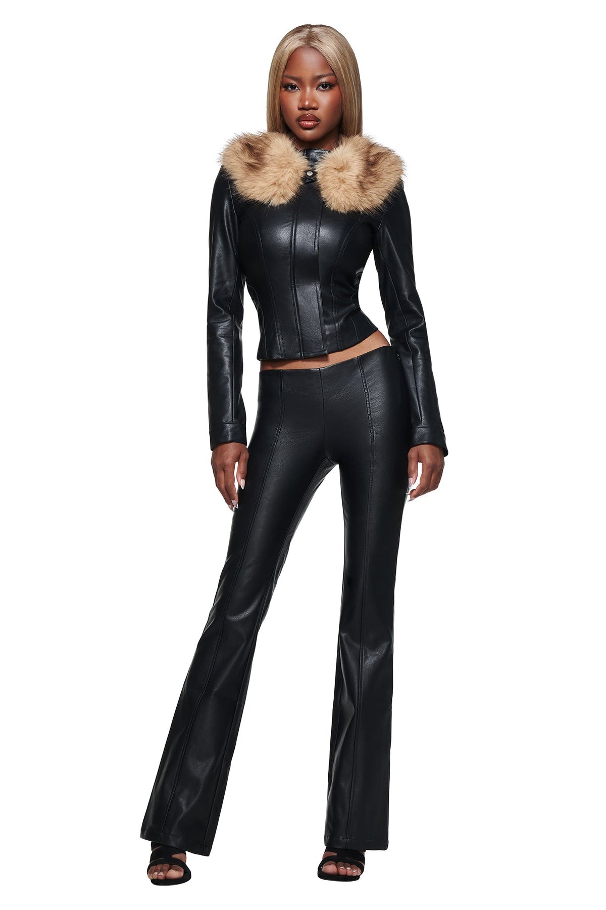 A woman stands against a white background, wearing the I.AM.GIA DUA PU PANT in black—faux leather pants detailed with decorative piping—paired with a black leather jacket featuring a tan fur collar. She has straight, blonde hair and completes her look with black heeled sandals.