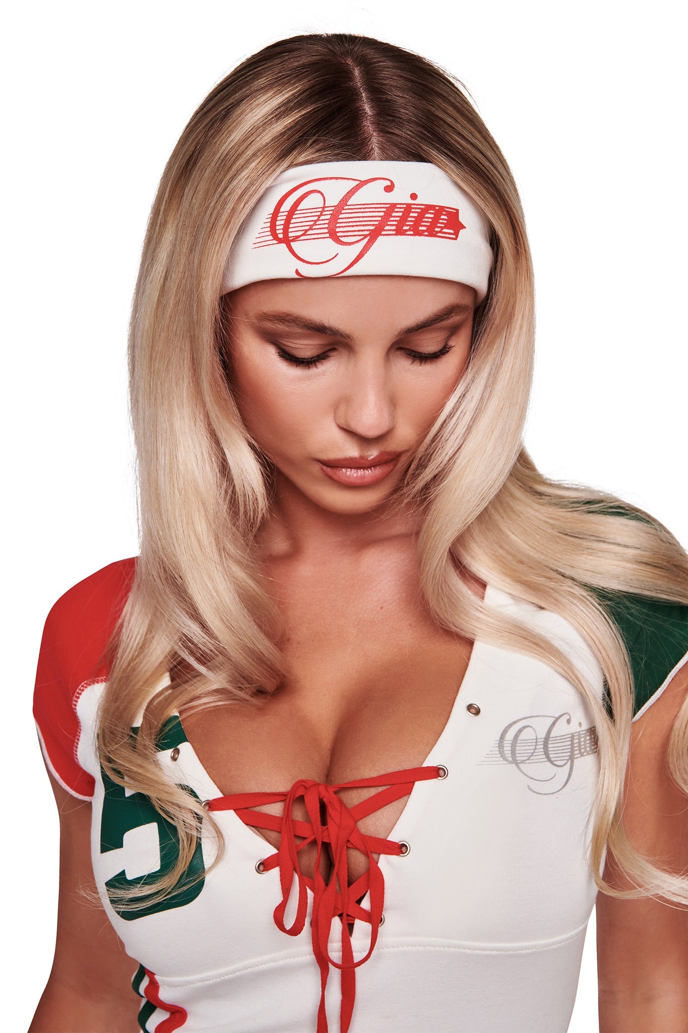 A woman with long blonde hair looks down, wearing the NOVIA SPORT HEADBAND in white by I.AM.GIA, featuring red script text. She pairs it with a coordinating lace-up top in white, red, and green with metallic studs and matching script.