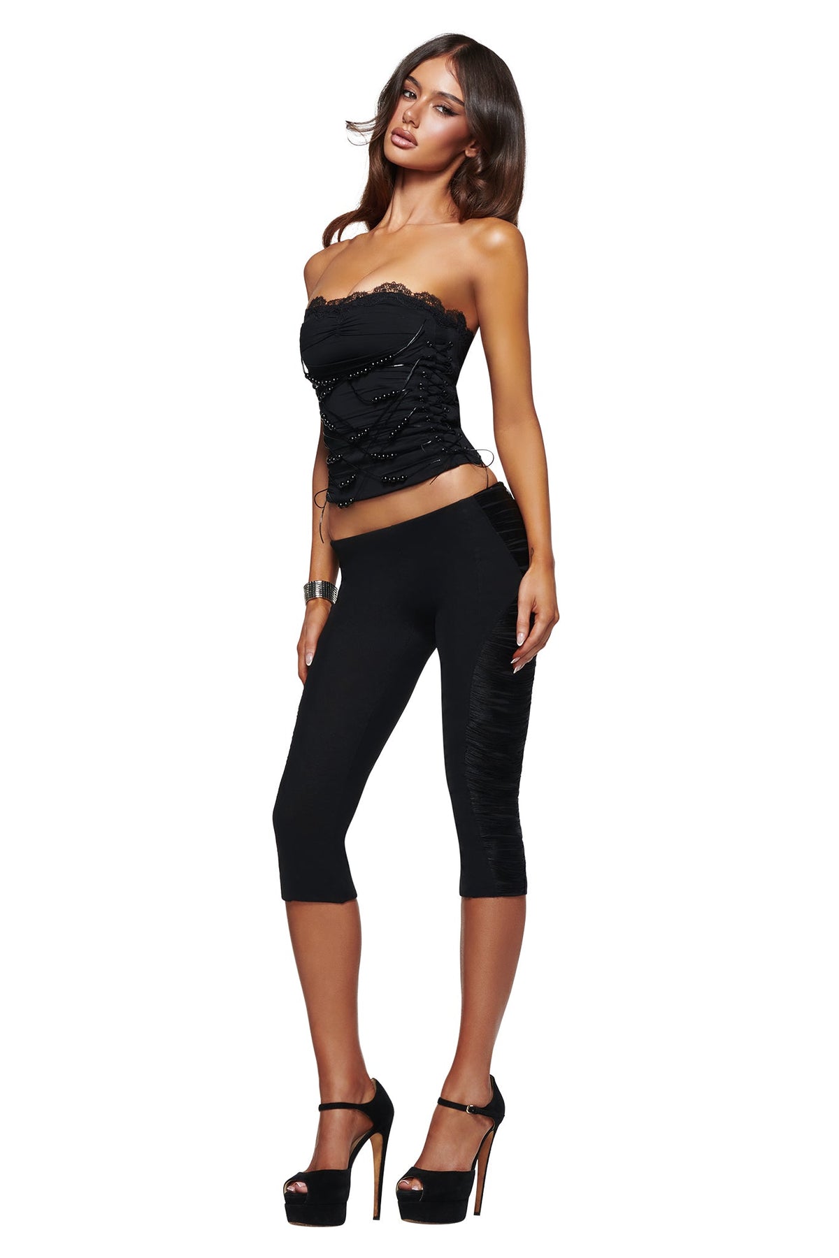 A woman wearing the I.AM.GIA DIXI CAPRI - BLACK bodycon capri leggings and a black strapless lace-detailed top, standing in high heels against a white background.