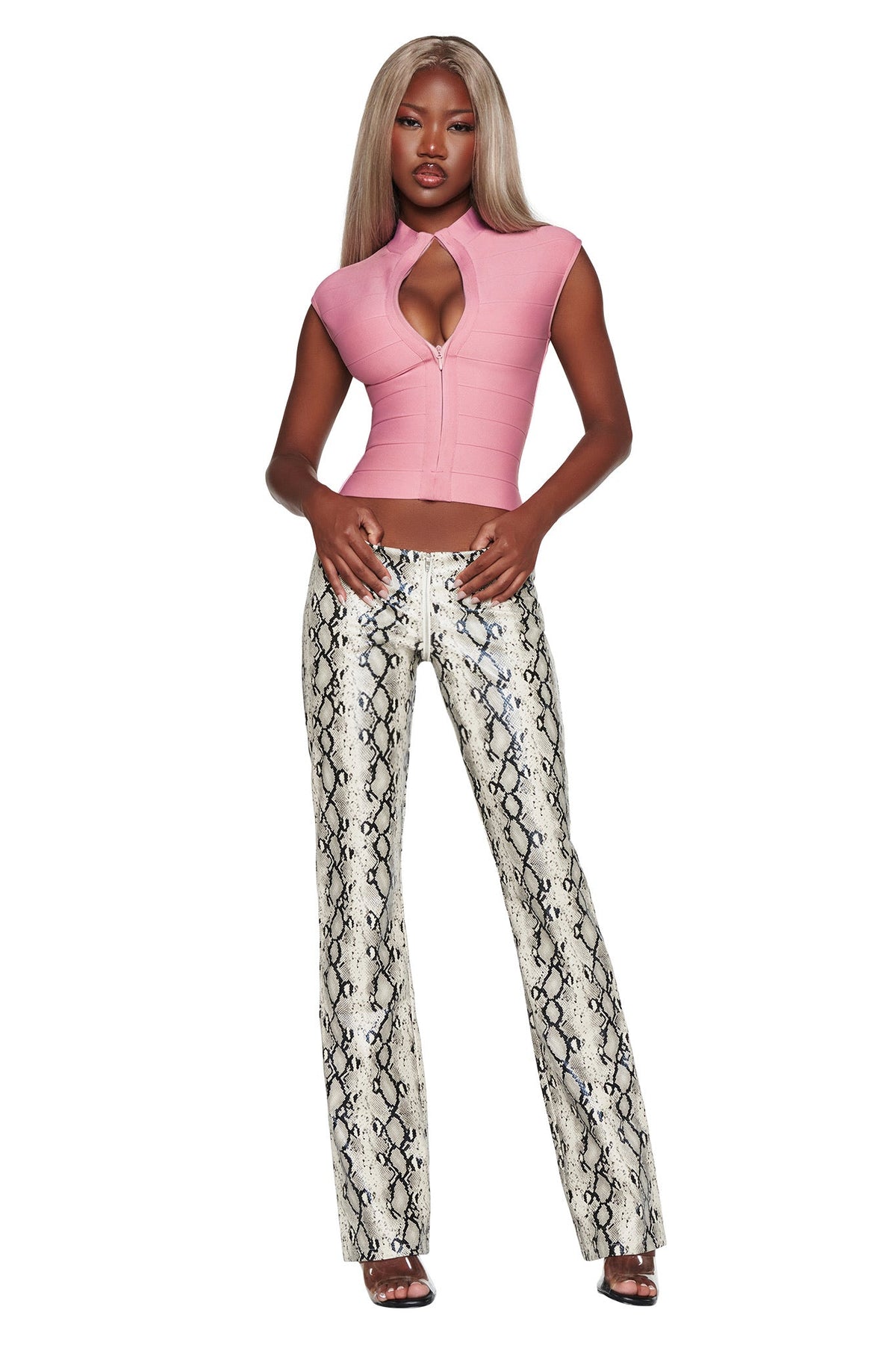 A woman stands wearing the I.AM.GIA ISELA BANDAGE TOP in dusty rose, a short-sleeve crop top featuring a front cutout, paired with fitted snakeskin-print pants. She has long, straight blonde hair and black heels.