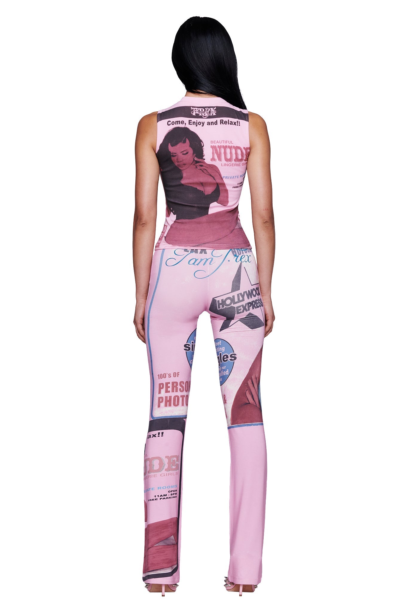 A woman stands facing away, wearing the T-REX COURTNEY TANK in pink by I.AM.GIA—a sleeveless top featuring retro-style graphics and phrases like "Come, Enjoy and Relax!" and "NUDE," with bold prints of women. Her outfit channels the playful, statement-making style of a baby pink graphic muscle tank.