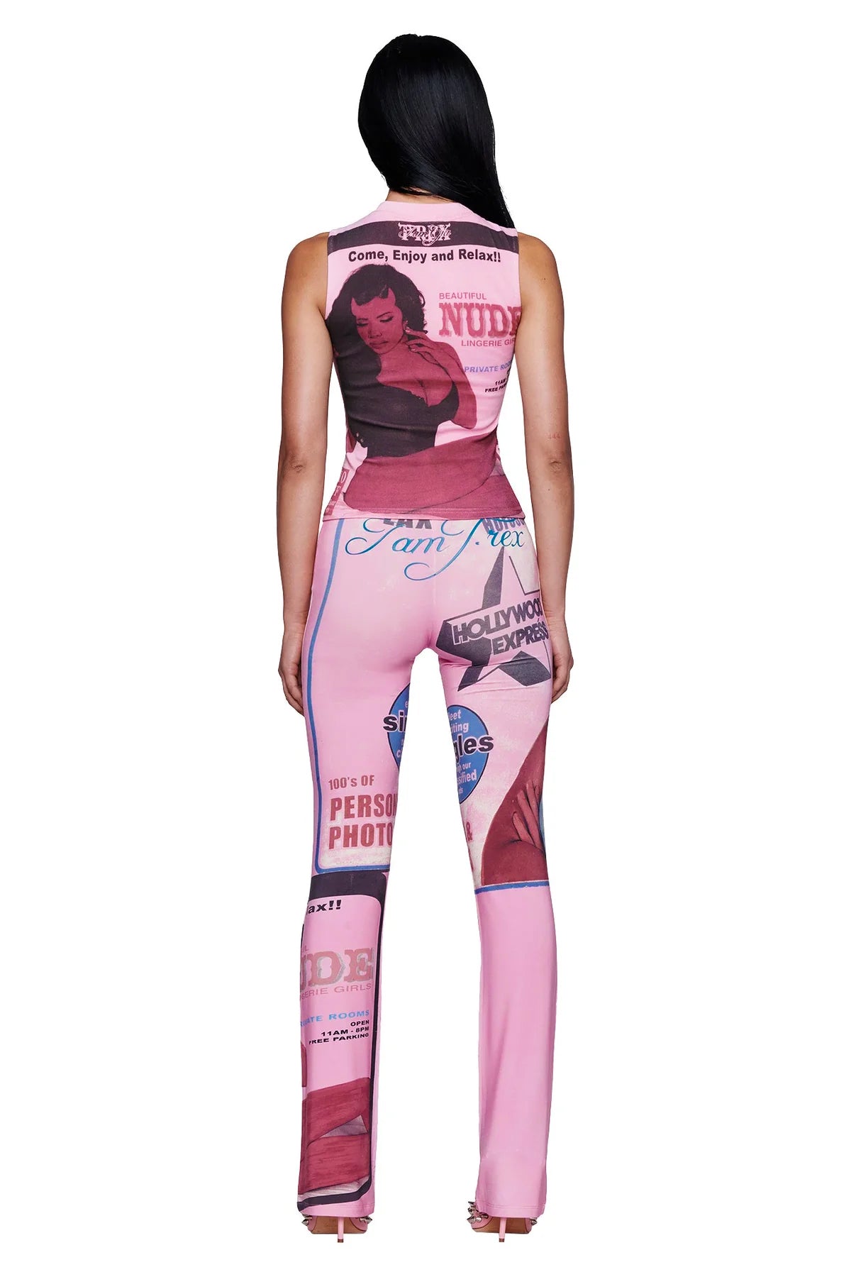 A woman stands facing away, wearing the I.AM.GIA T-REX COURTNEY TANK in pink—a sleeveless jumpsuit adorned with exclusive vintage pin-up and magazine cover placement prints—paired with open-toe heels. Her long, dark hair cascades down her back.