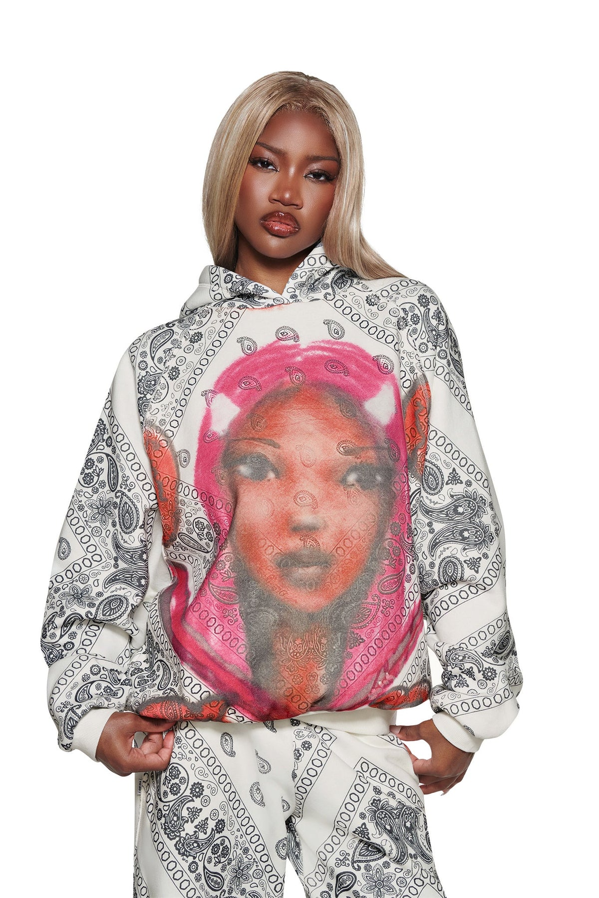A woman with long blonde hair wears the I.AM.GIA T-REX PULLOVER HOODIE - PAISLEY FACE, a boxy fleece hoodie with black paisley patterns and a bold pink portrait print, standing confidently against white.