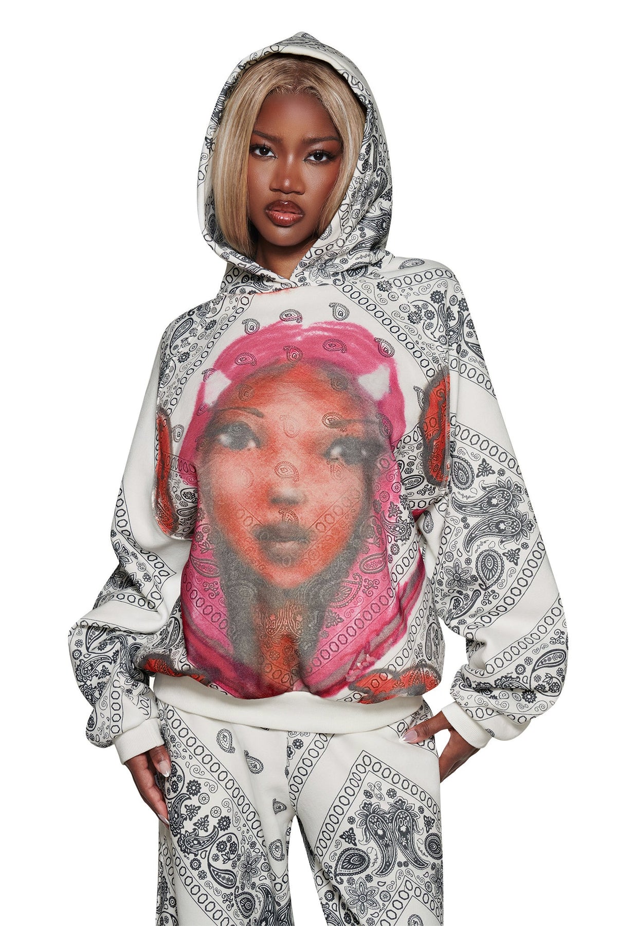 Wearing the I.AM.GIA T-REX PULLOVER HOODIE - PAISLEY FACE, which features black paisley patterns and a colorful graphic of a woman’s face, a person stands with one hand on their hip.