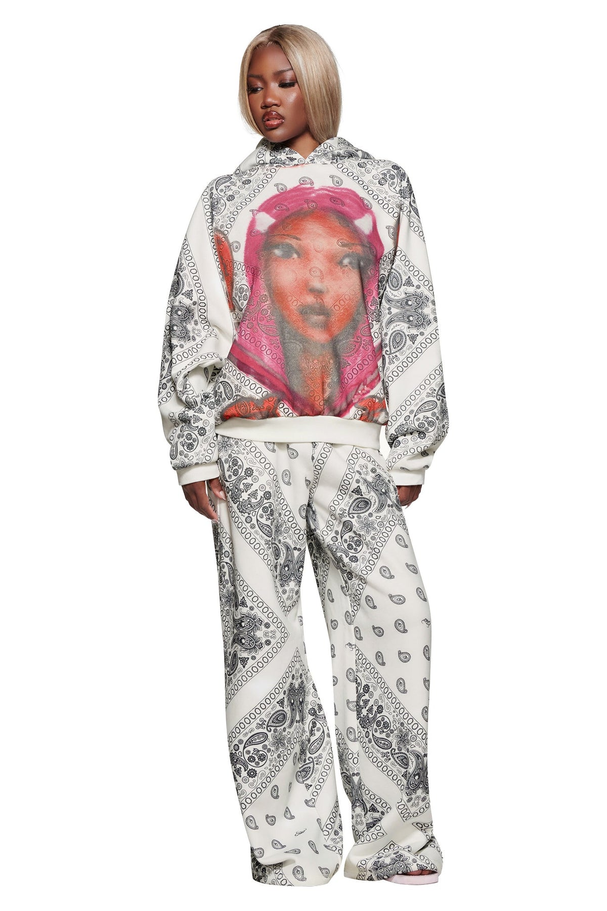 A person wears the I.AM.GIA T-REX PULLOVER HOODIE - PAISLEY FACE, a plush fleece bandana-print hoodie with oversized hood and pink-hued woman’s portrait, paired with matching pants against a white background.