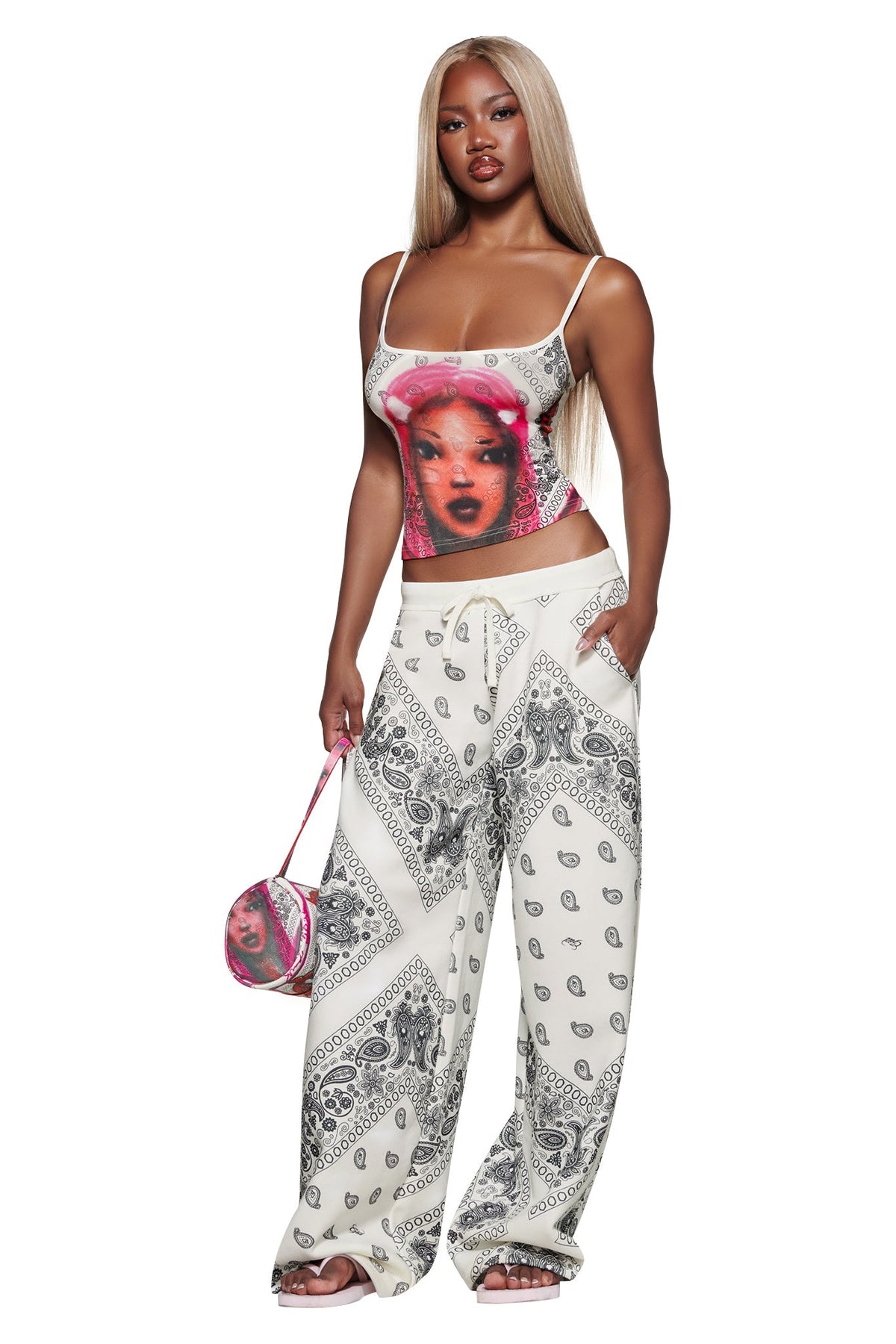 A woman with long blonde hair wears a colorful graphic tank, I.AM.GIA T-REX TRACKPANT - PAISLEY, and carries a small pink patterned bag, standing facing forward against a white background.