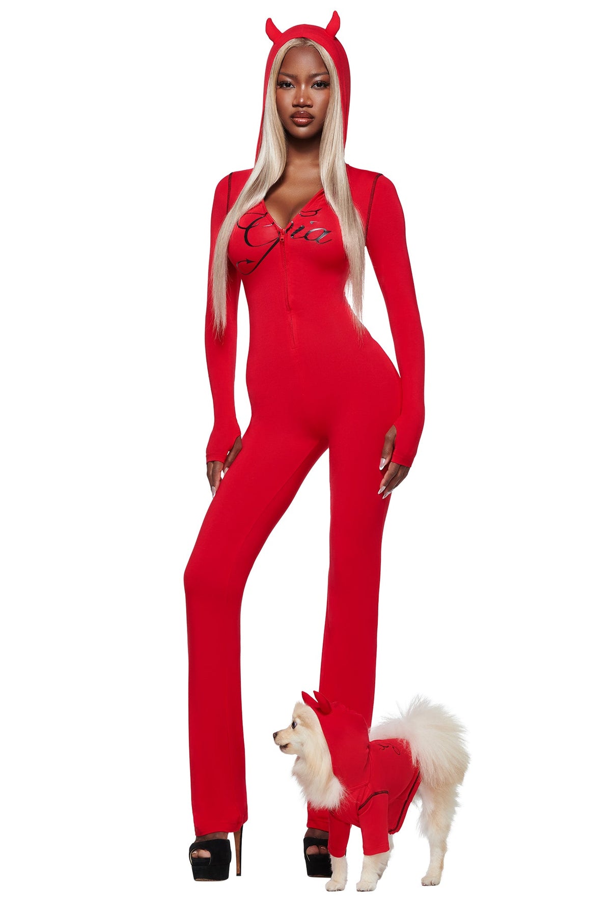 A woman wears the I.AM.GIA BLARE HORN JUMPSUIT - RED with a devil horn hood, confidently posing next to her small dog in a matching outfit against a white background.