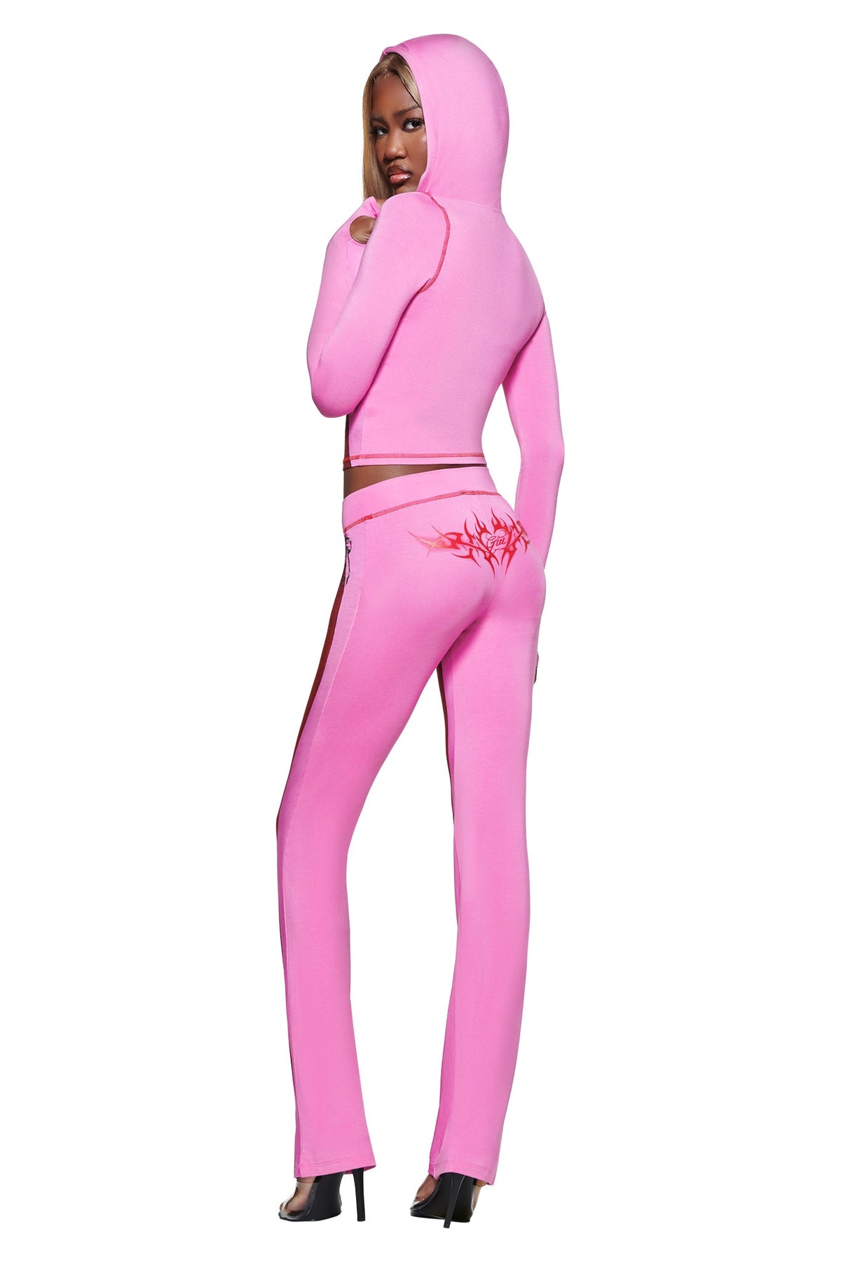 Woman in the I.AM.GIA T-REX SPRAY BODY BLARE TRACKPANT - PINK and matching pink hooded crop sweatshirt, with a red back graphic, looks over her shoulder while standing in heeled sandals.