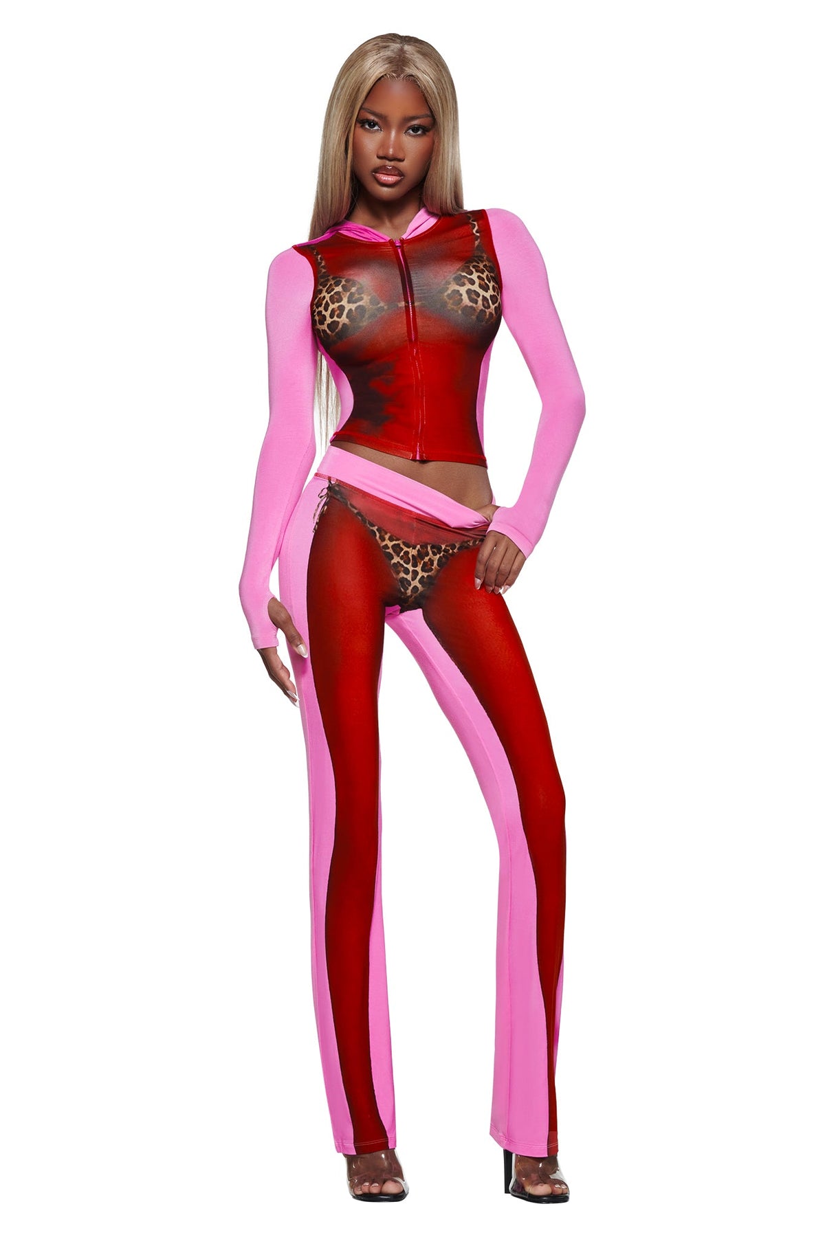A woman with long blonde hair wears the T-REX SPRAY BODY BLARE TRACKPANT - PINK by I.AM.GIA, featuring an airbrushed bikini print and leopard details, paired with black heels against a white background.