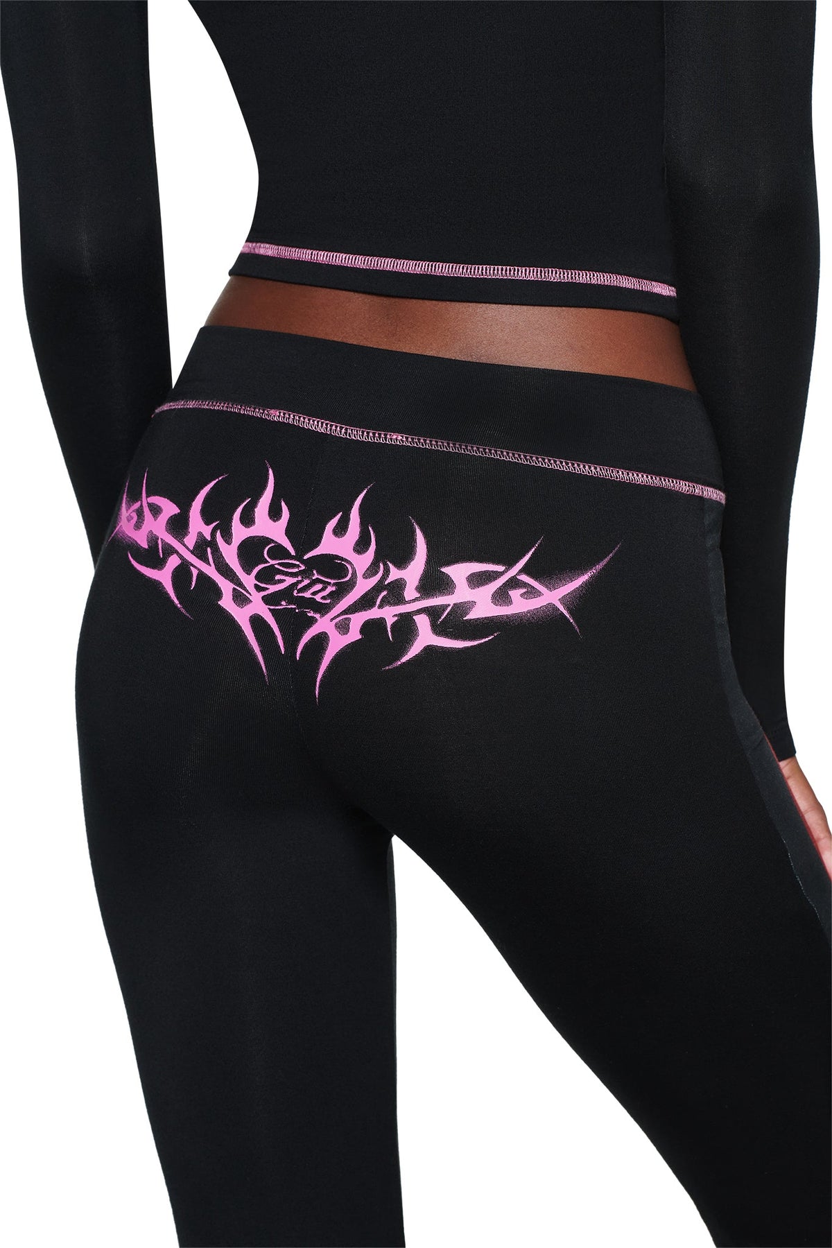 A person wearing T-REX SPRAY BODY BLARE TRACKPANT - BLACK by I.AM.GIA with pink tribal flames and “Girl” on the back, shown waist down, perfect for airport style against a white background.