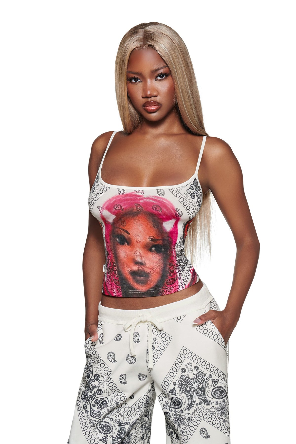 A woman with long blonde hair wears the I.AM.GIA T-REX TANK - PAISLEY FACE, a white crop camisole with a colorful face graphic, paired with paisley pants and standing against a white background.