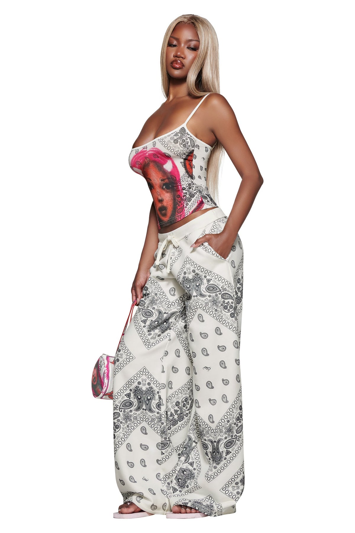 Woman with long blonde hair wears the I.AM.GIA T-REX TANK - PAISLEY FACE, wide-leg paisley pants, sandals, and holds a small matching purse while posing with one hand in her pocket against a white background.