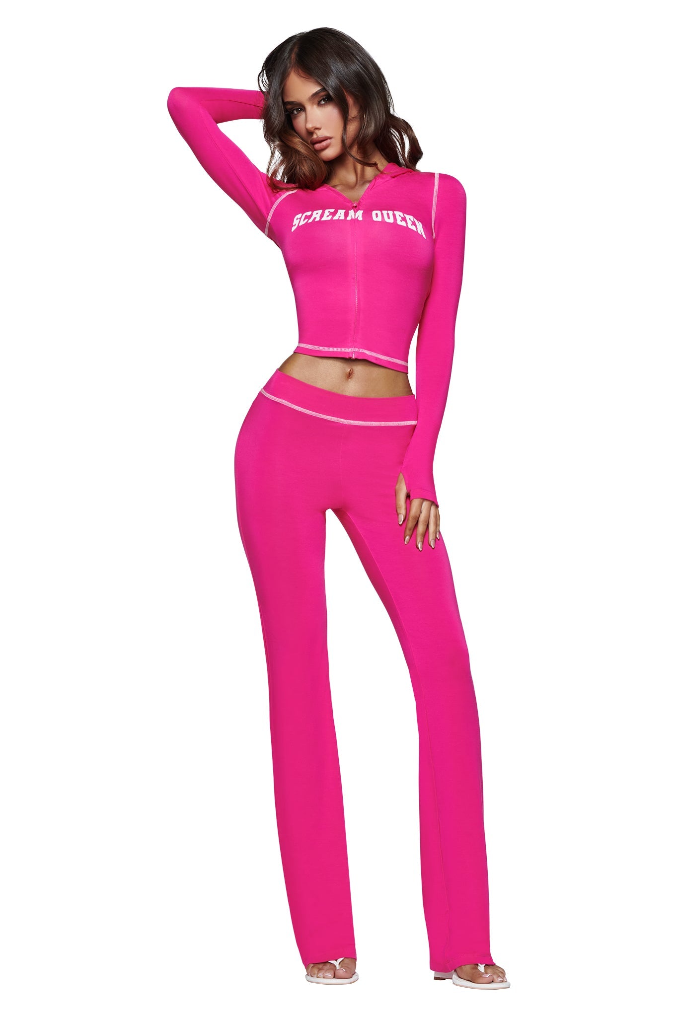 A woman wearing the I.AM.GIA BLARE HOODIE in hot pink Scream Queen, paired with matching high-waisted pants and clear high-heeled shoes, poses with one hand on her head.