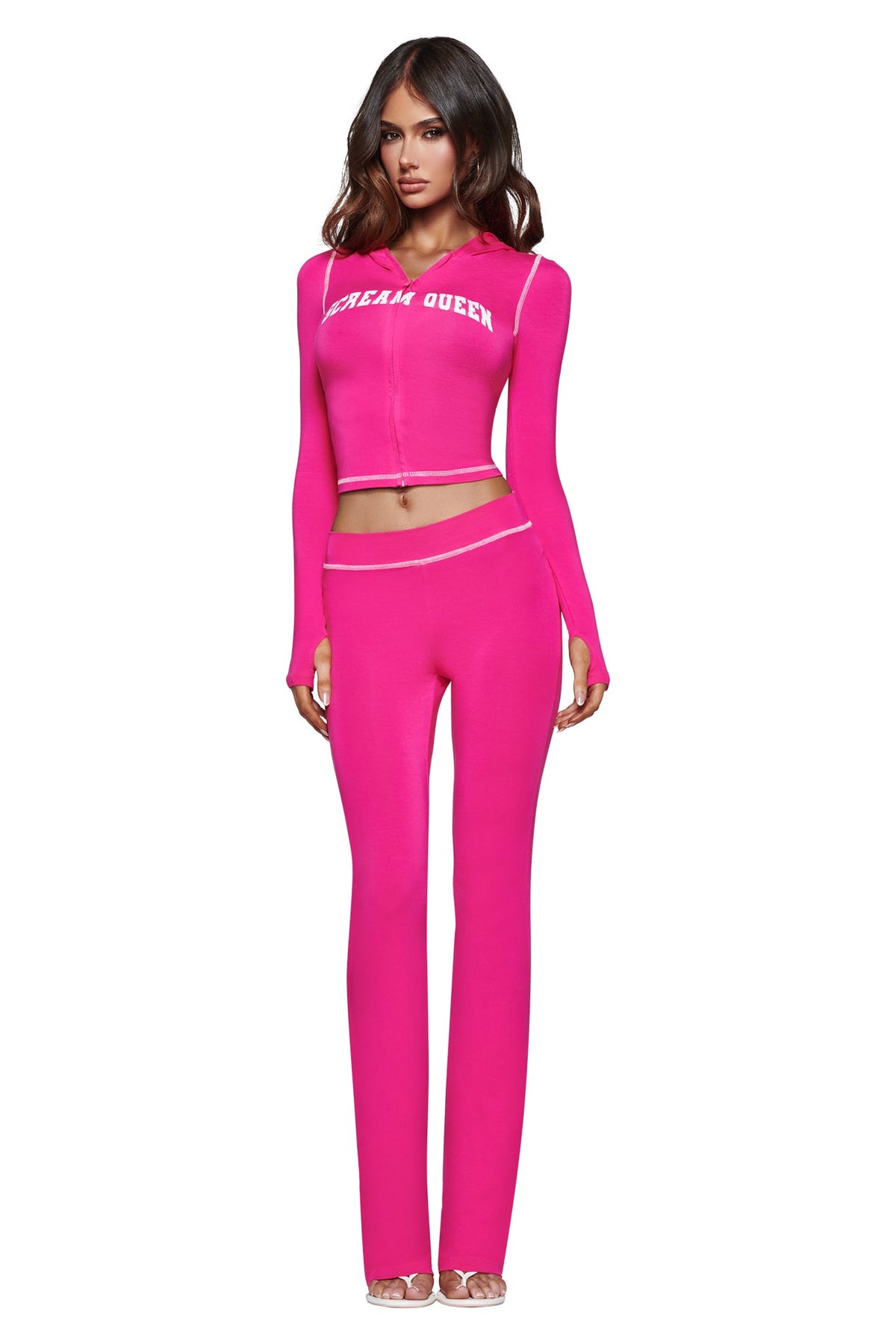 A woman stands against a white background, wearing the I.AM.GIA BLARE TRACKPANT - HOT PINK SCREAM QUEEN set, featuring a hot pink, long-sleeve crop top with a bold Scream Queen graphic and matching high-waisted trackpants, paired with clear high-heeled shoes.