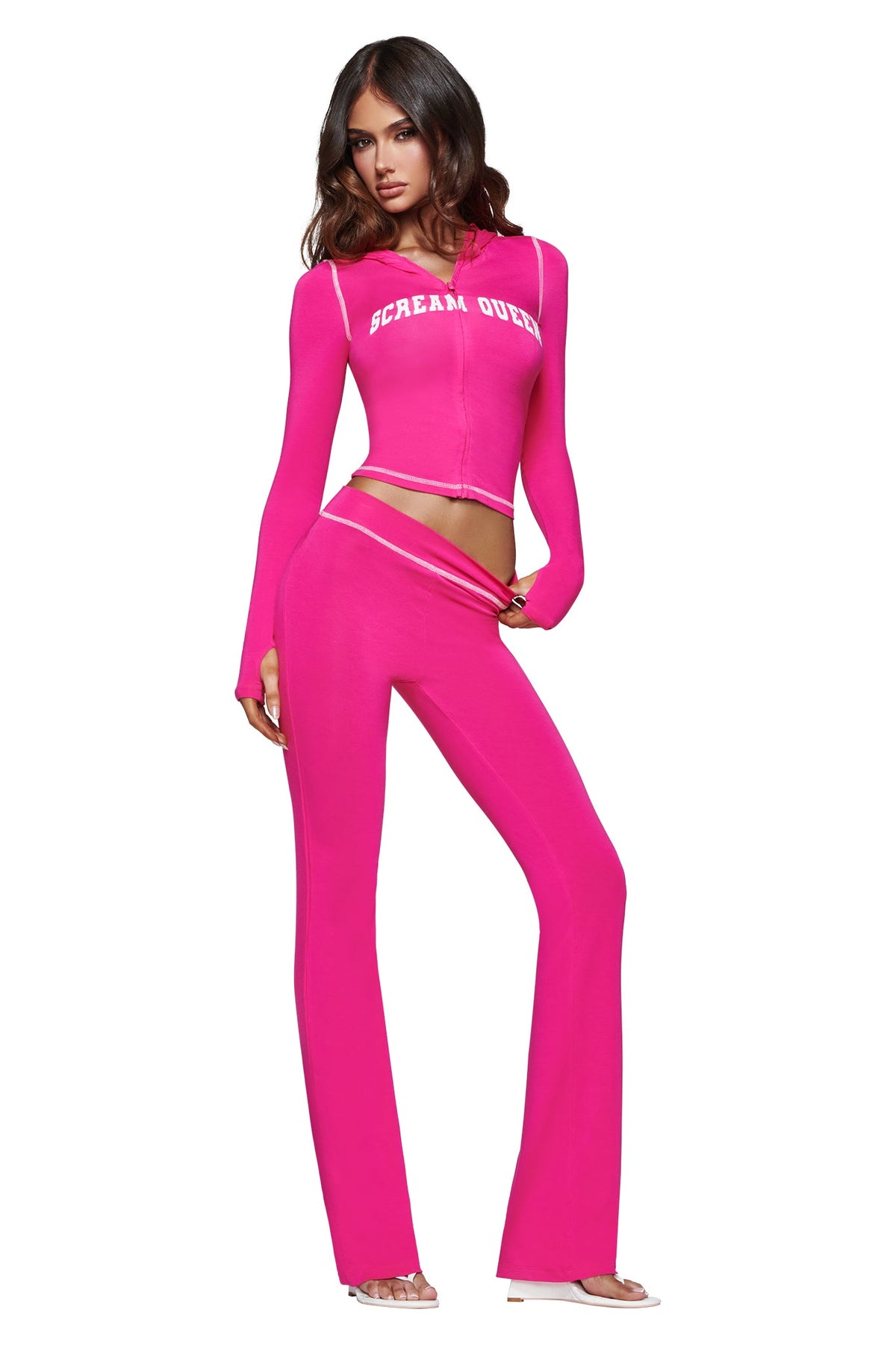 A woman poses in the I.AM.GIA BLARE HOODIE - HOT PINK SCREAM QUEEN, a vibrant pink cropped hoodie featuring a bold “Scream Queen” graphic. She pairs it with matching flared pants and clear high-heeled sandals, standing against a white background.