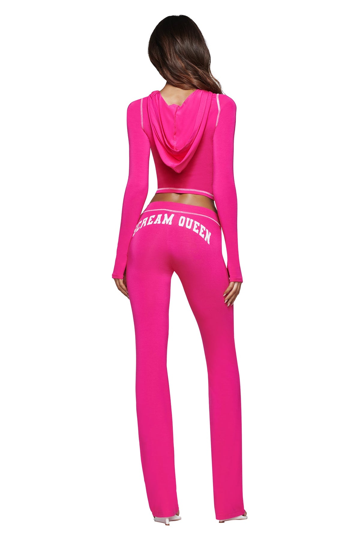 A woman stands with her back to the camera, dressed in the I.AM.GIA BLARE TRACKPANT – HOT PINK SCREAM QUEEN, a vibrant hot pink hooded tracksuit featuring “DREAM QUEEN” in white lettering on the back of the pants and a striking Scream Queen graphic.