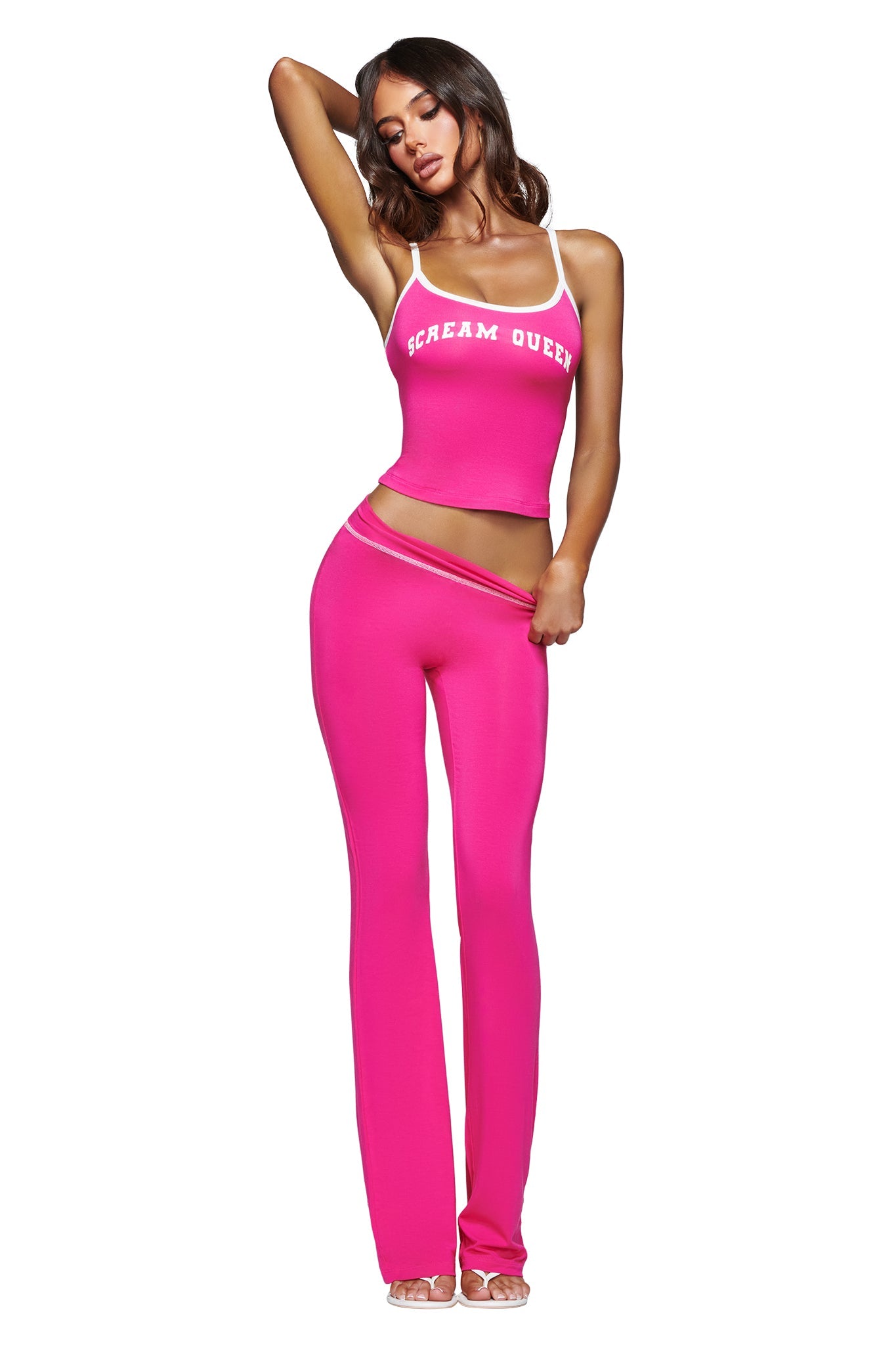 A woman poses in a vibrant look, wearing the I.AM.GIA BLARE SINGLET in Hot Pink SCREAM QUEEN, paired with matching high-waisted pants. She completes the outfit with clear high-heeled shoes, striking a confident pose with one arm raised and the other on her waistband. The bold BLARE SINGLET serves as the standout piece, adding an edgy statement to her ensemble.