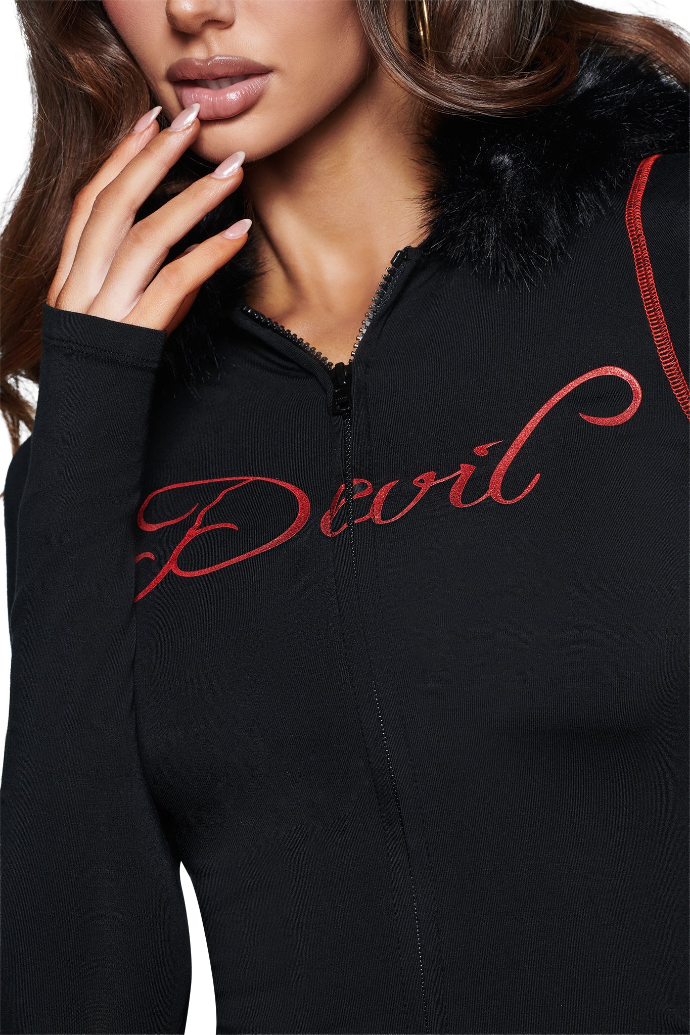 A woman wears the I.AM.GIA BLARE FUR TRIM HOODIE in black, featuring a zip-up front, luxe black fur collar, red piping on the shoulders, and “Devil” boldly scripted in red across the chest. The edgy hoodie adds to her statement look as she touches her face, accentuating her long nails.