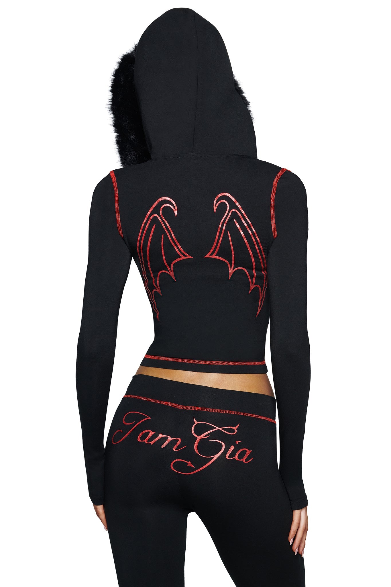 A person is seen from behind wearing the I.AM.GIA BLARE FUR TRIM HOODIE in black, featuring dramatic red bat wings on the back and a bold devil graphic design. The hood is up, and “Tam Gia” is scripted across the back of their pants.