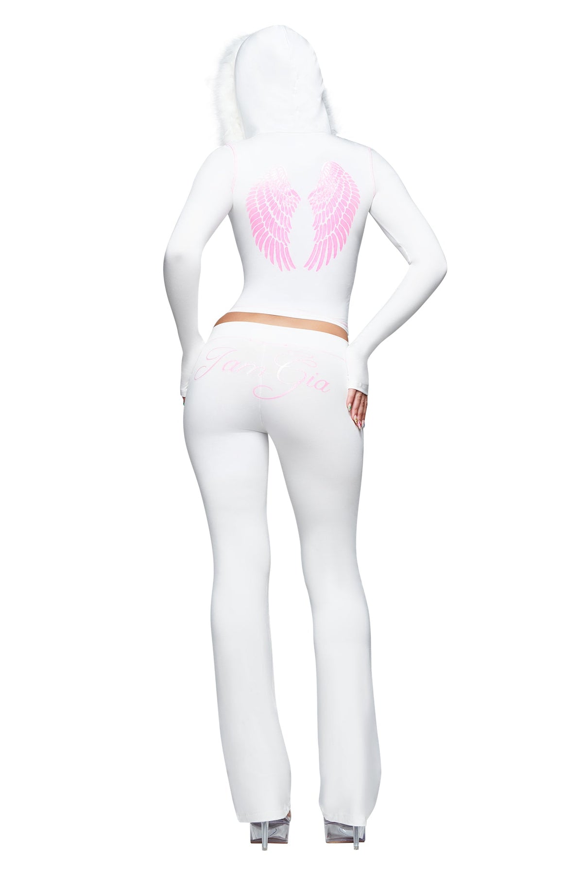 A person stands with one hand on their hip and high heels, photographed from behind, wearing the I.AM.GIA BLARE TRACKPANT - WHITE : ANGEL. This bamboo trackpant features a white hooded outfit with pink angel wings graphic on the back, text detailing on the pants, a low-rise waist, and sleek flared legs for a modern silhouette.