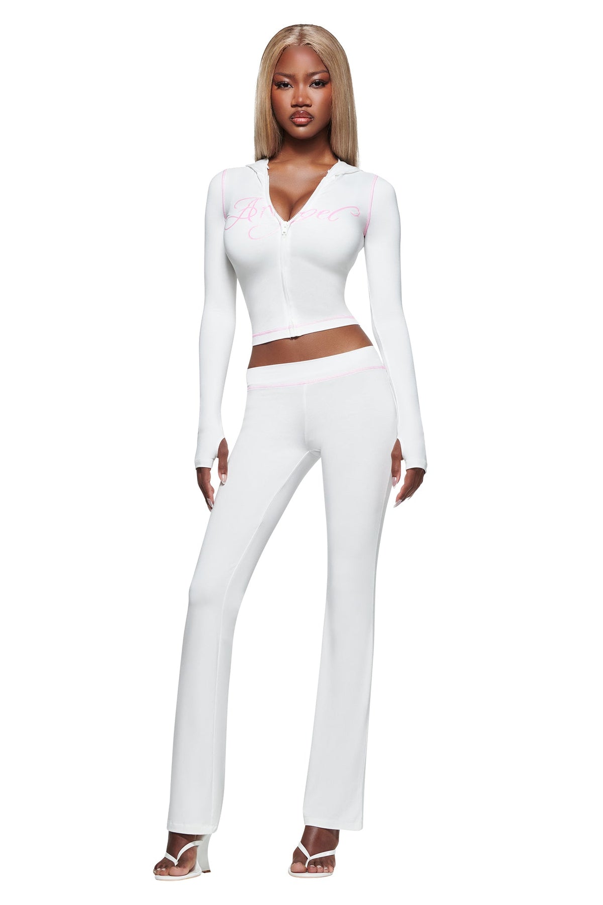 A woman stands posing against a plain white background, wearing the I.AM.GIA BLARE TRACKPANT - WHITE : ANGEL with a low-rise waist and flared legs, styled with a fitted, long-sleeve white zip-up top and white heeled sandals.