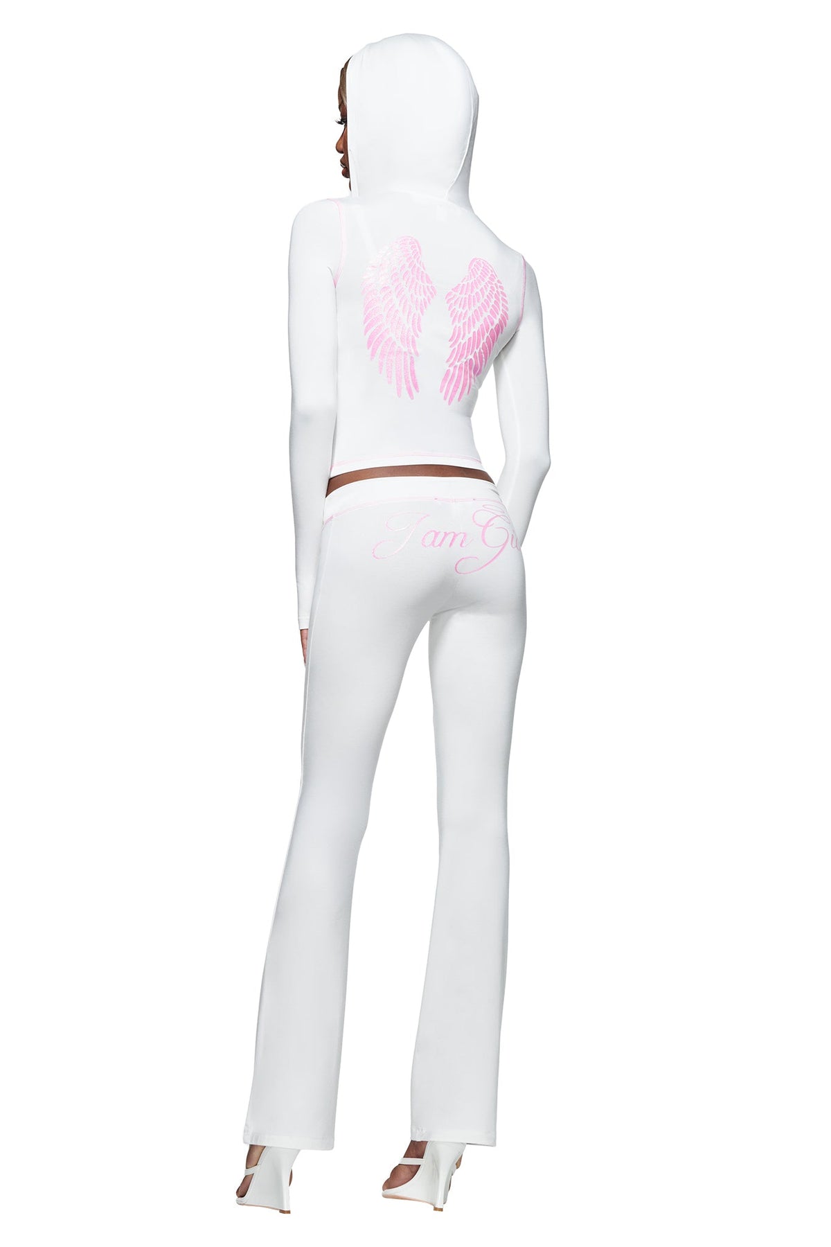 Wearing the I.AM.GIA BLARE TRACKPANT - WHITE : ANGEL, which features pink angel wings on the back and “I am G” printed on the low-rise, flared leg silhouette, a person stands facing away in a white hooded outfit paired with white high-heeled shoes.