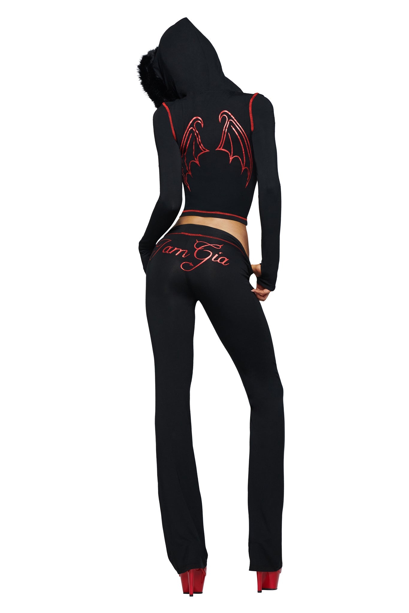 A person, seen from behind with hands on hips, sports the I.AM.GIA BLARE TRACKPANT - JET BLACK : DEVIL featuring a bold black devil GIA logo and the “I.AM.GIA” branding, paired with a black hooded outfit adorned with striking red bat wings and finished off with red high heels.