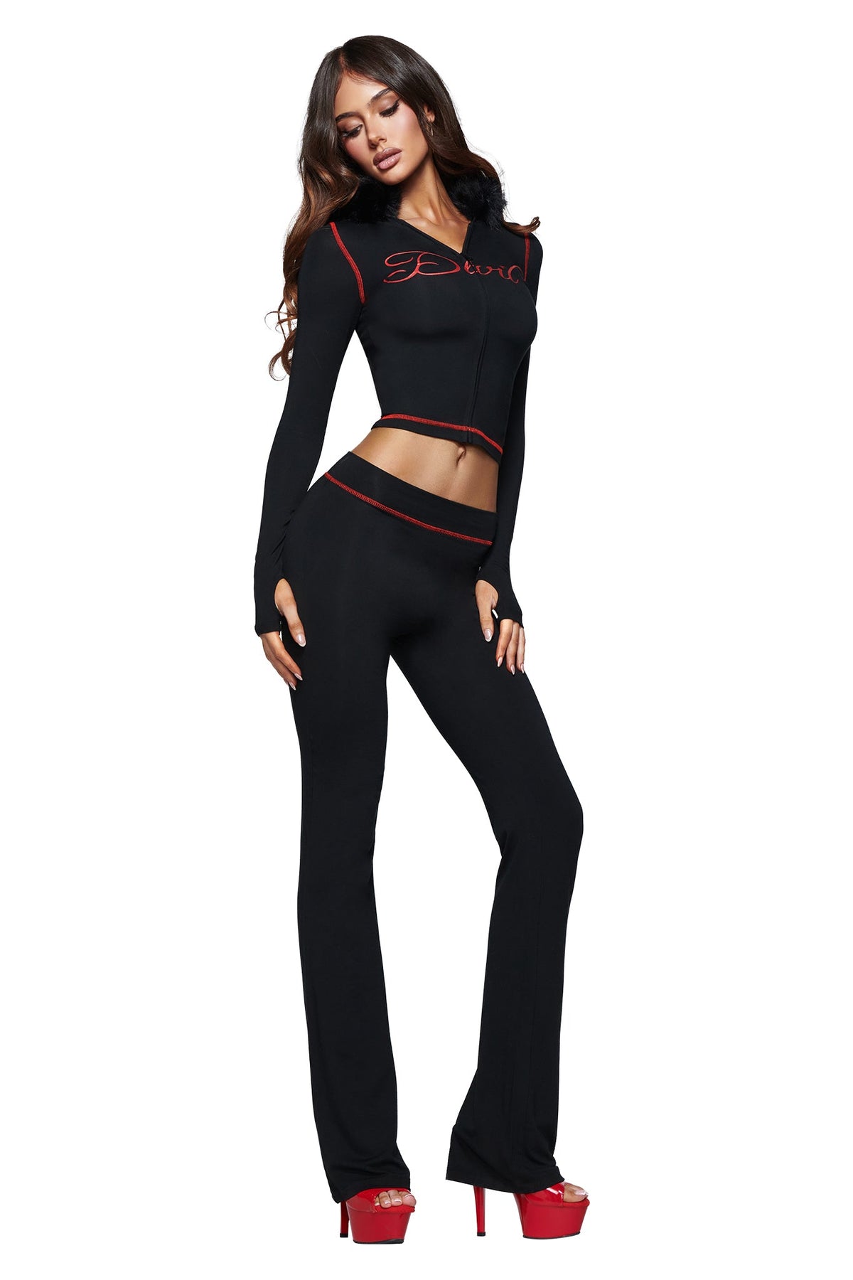 A woman poses in a black crop top and matching I.AM.GIA BLARE TRACKPANT - JET BLACK : DEVIL with red trim—crafted from soft bamboo fabrication—and red high-heeled platform shoes, standing against a white background. The outfit is finished with the signature black devil GIA logo detail.