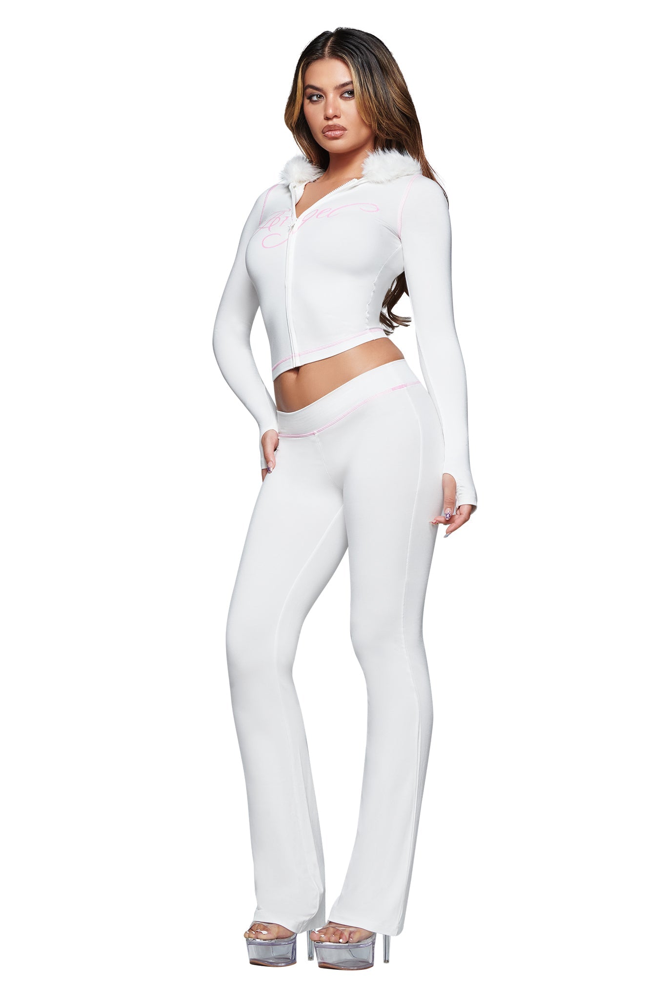A woman stands wearing I.AM.GIA’s BLARE TRACKPANT - WHITE : ANGEL, paired with a fitted white zip-up hoodie featuring a faux fur collar. She has long brown hair and finishes the look with high platform silver heels against a plain white background.
