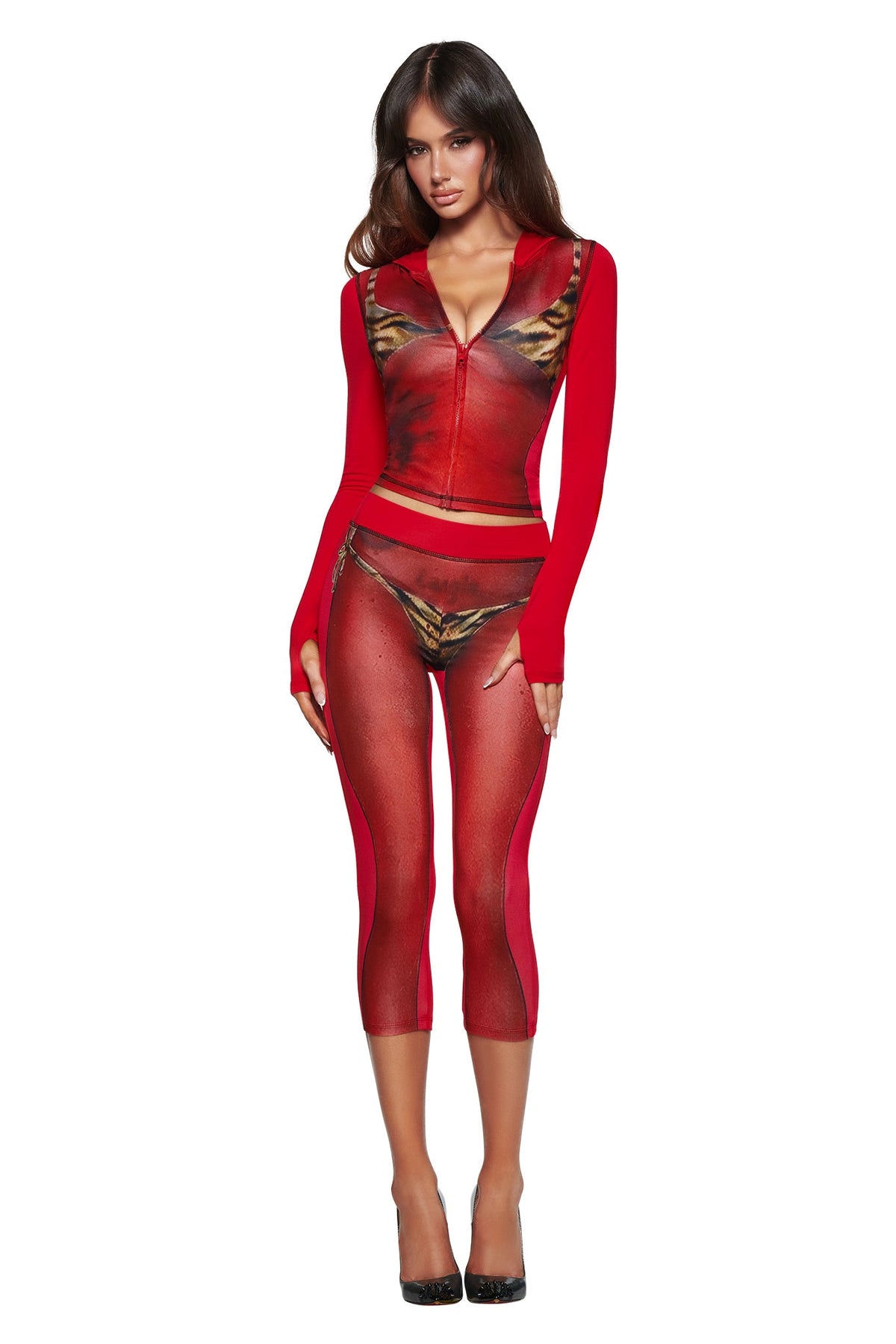 A woman wears the I.AM.GIA T-REX SPRAY BODY BLARE CAPRI - RED, featuring a red zip-up crop top and matching sheer red capri leggings with bikini body print, paired with black heels against a white background.