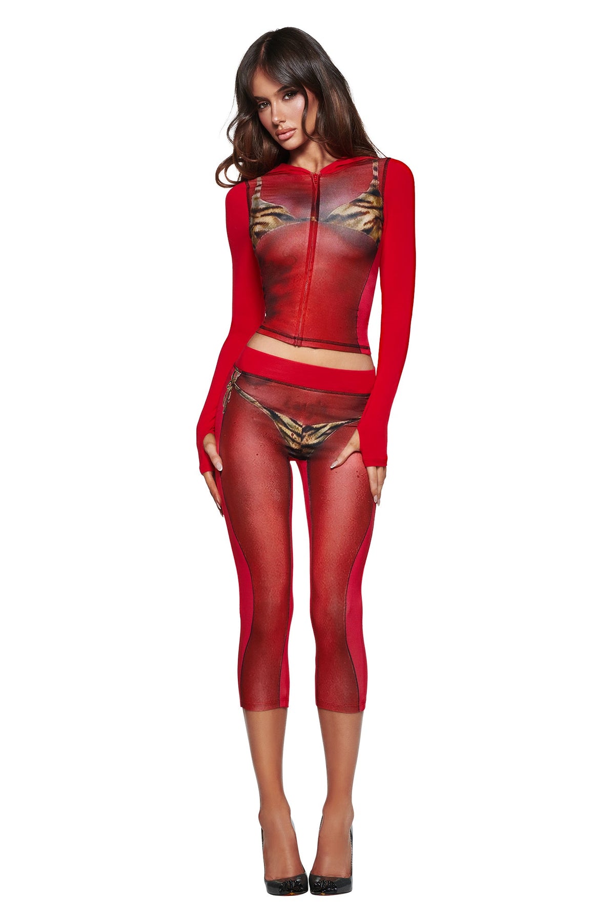 A woman stands facing forward in the I.AM.GIA T-REX SPRAY BODY BLARE CAPRI - RED, a form-fitting set with long sleeves and capri leggings, finished with black high heels.
