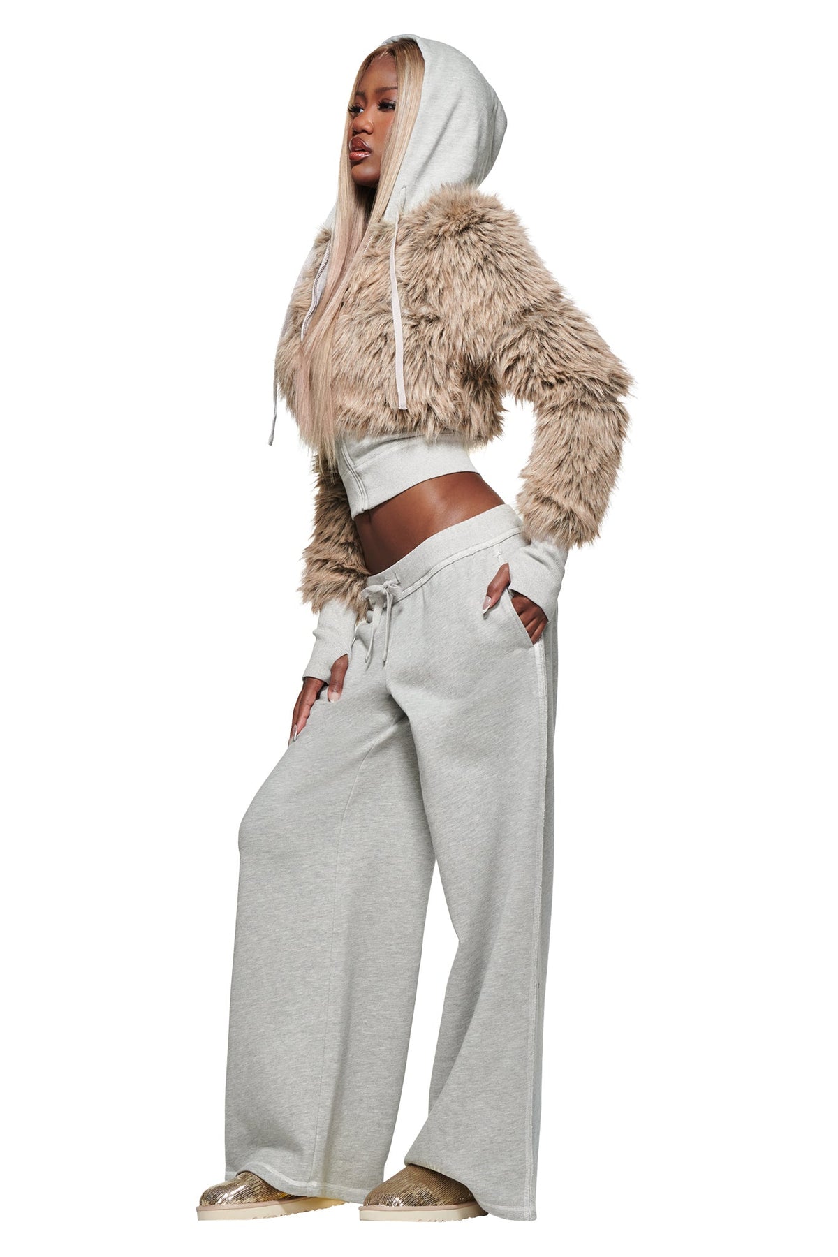 A woman with long, straight blonde hair stands against a white background, looking to the side with one hand on her hip. She wears loose KOKO TRACKPANT - HEATHER GREY by I.AM.GIA, paired with a beige furry cropped hoodie and light brown shoes.